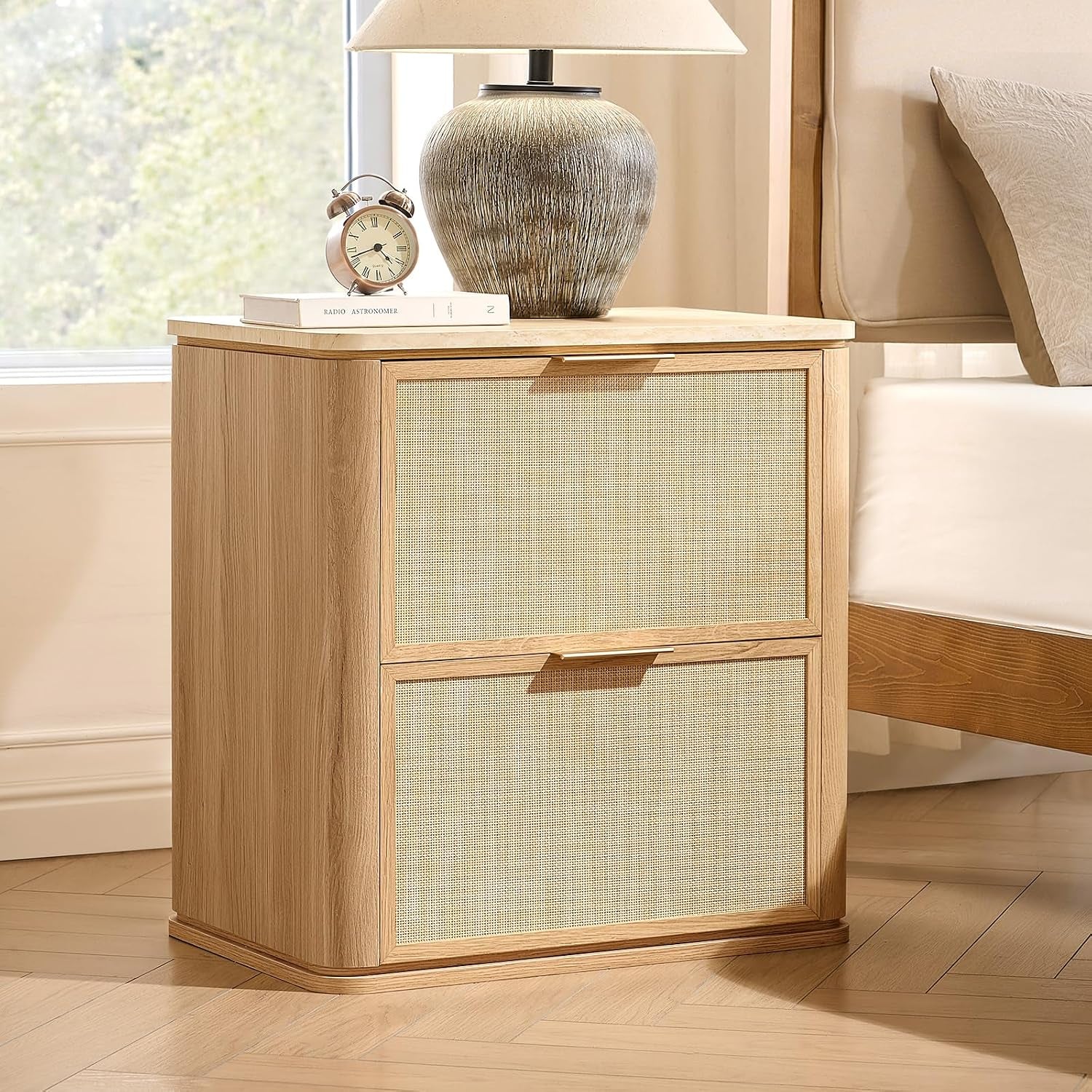 Rattan-look nightstand in natural oak beside a bed, front three-quarter view showing woven drawer fronts