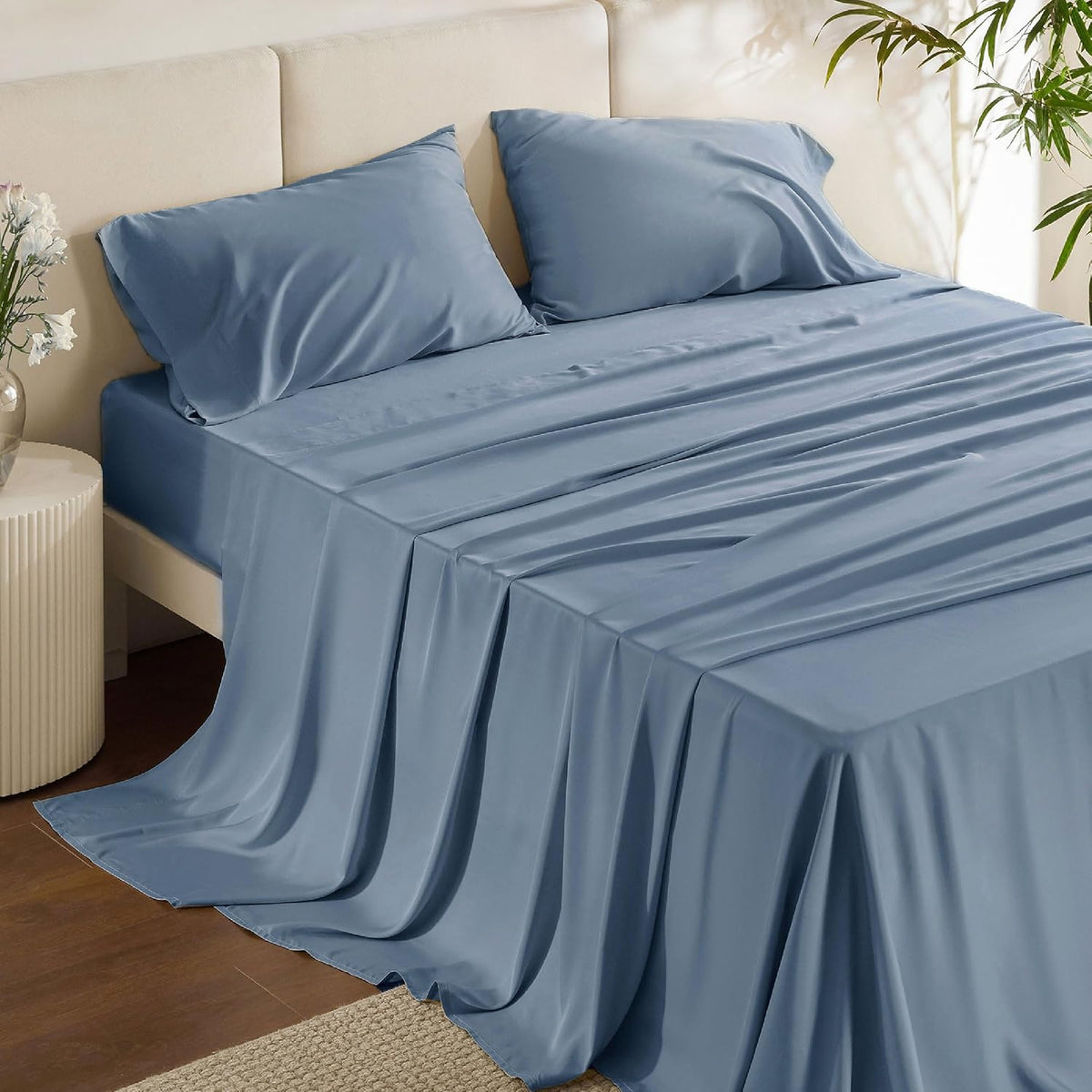 Three-quarter view of a made bed with medium blue silky sheets cascading to the floor and two pillows at the headboard.