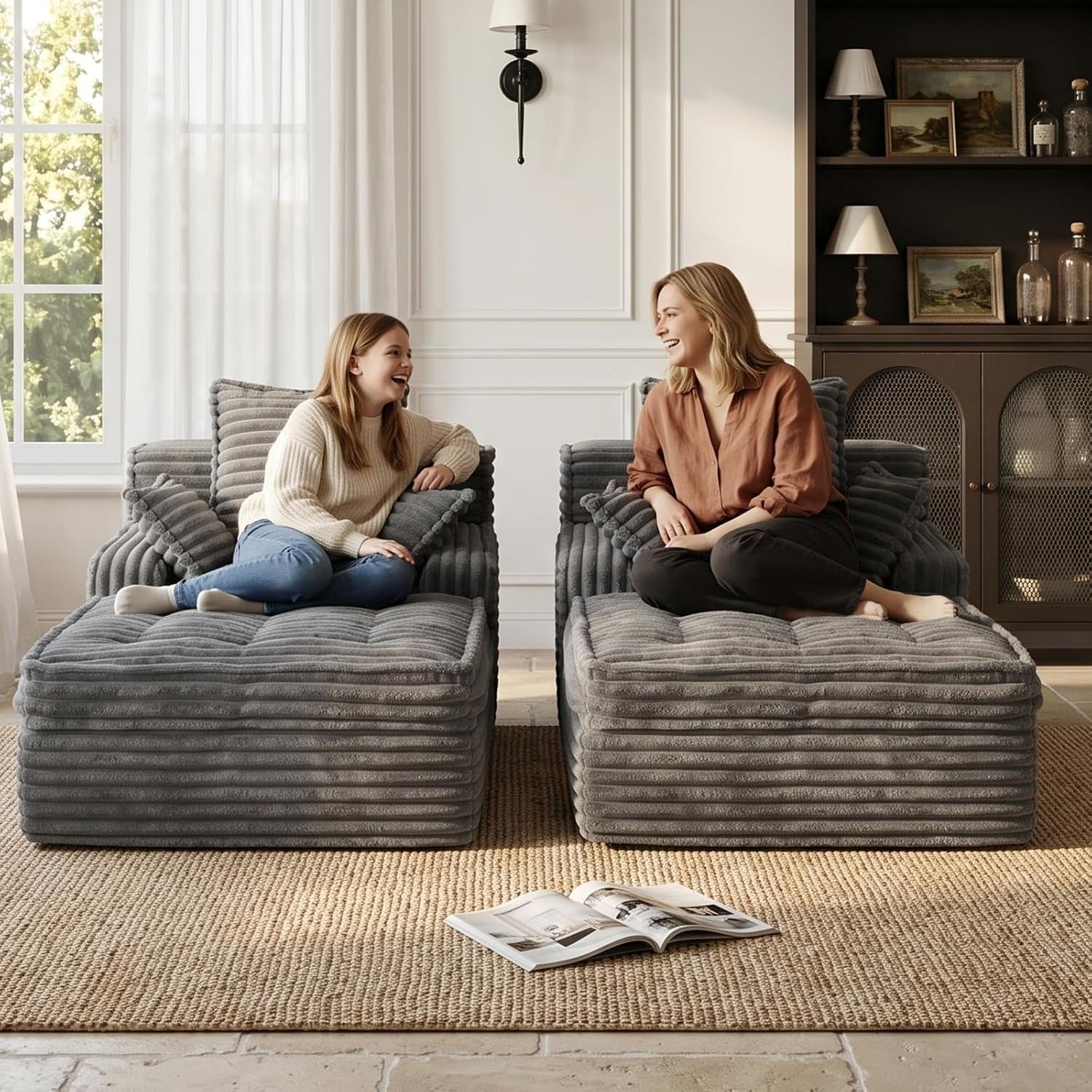 Two people laughing while seated on two matching gray chaise lounge sections in a bright living room.