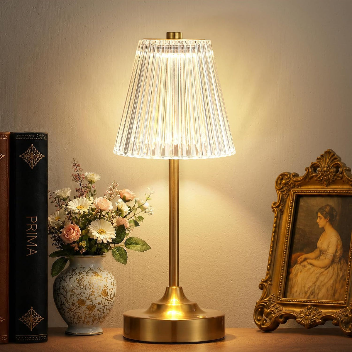 Gold table lamp lit on a bedside table with a clear pleated acrylic shade, vase of flowers and framed portrait