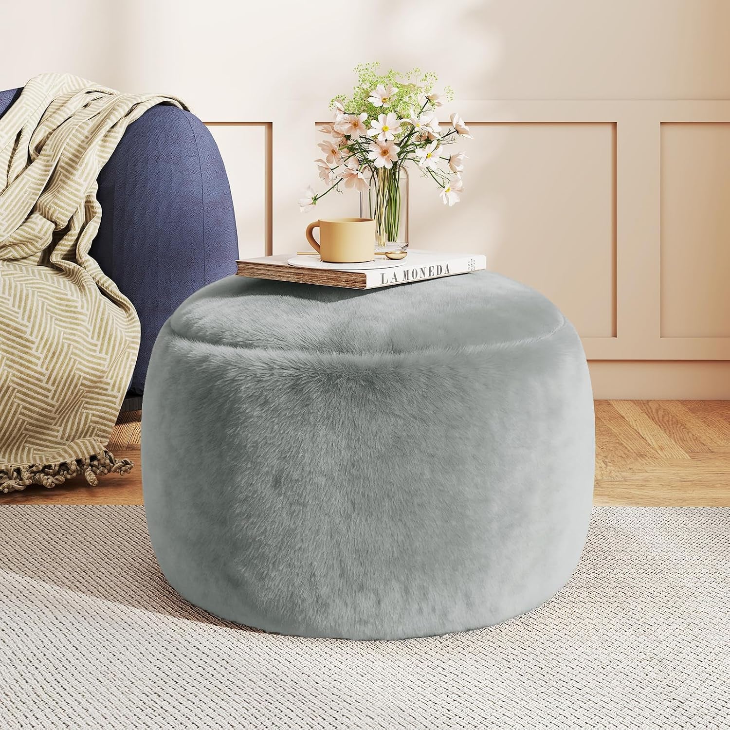 Grey round faux-fur pouf ottoman with book and vase on top beside a sofa