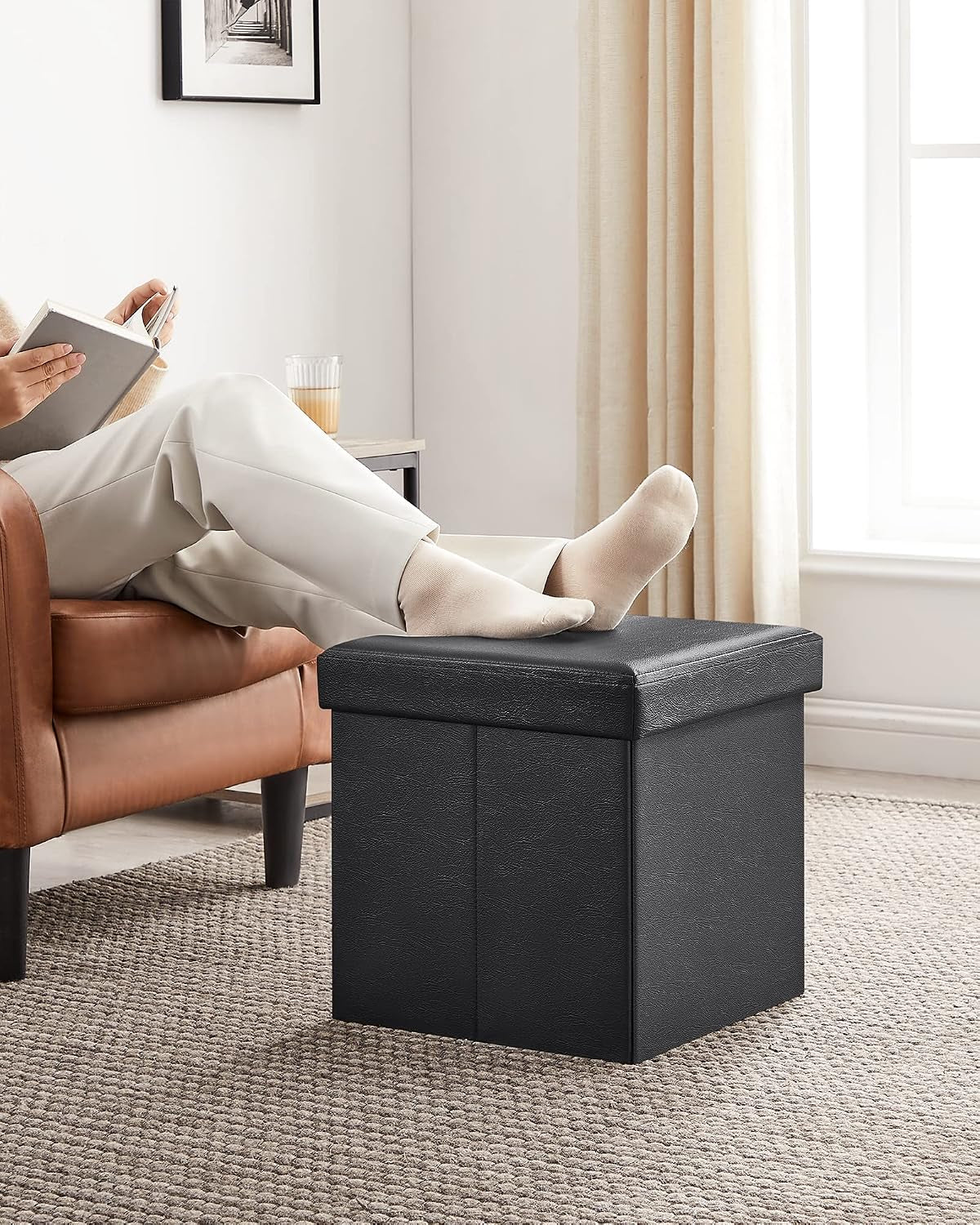 Black square storage ottoman placed on a carpet in front of an armchair with feet resting on top.
