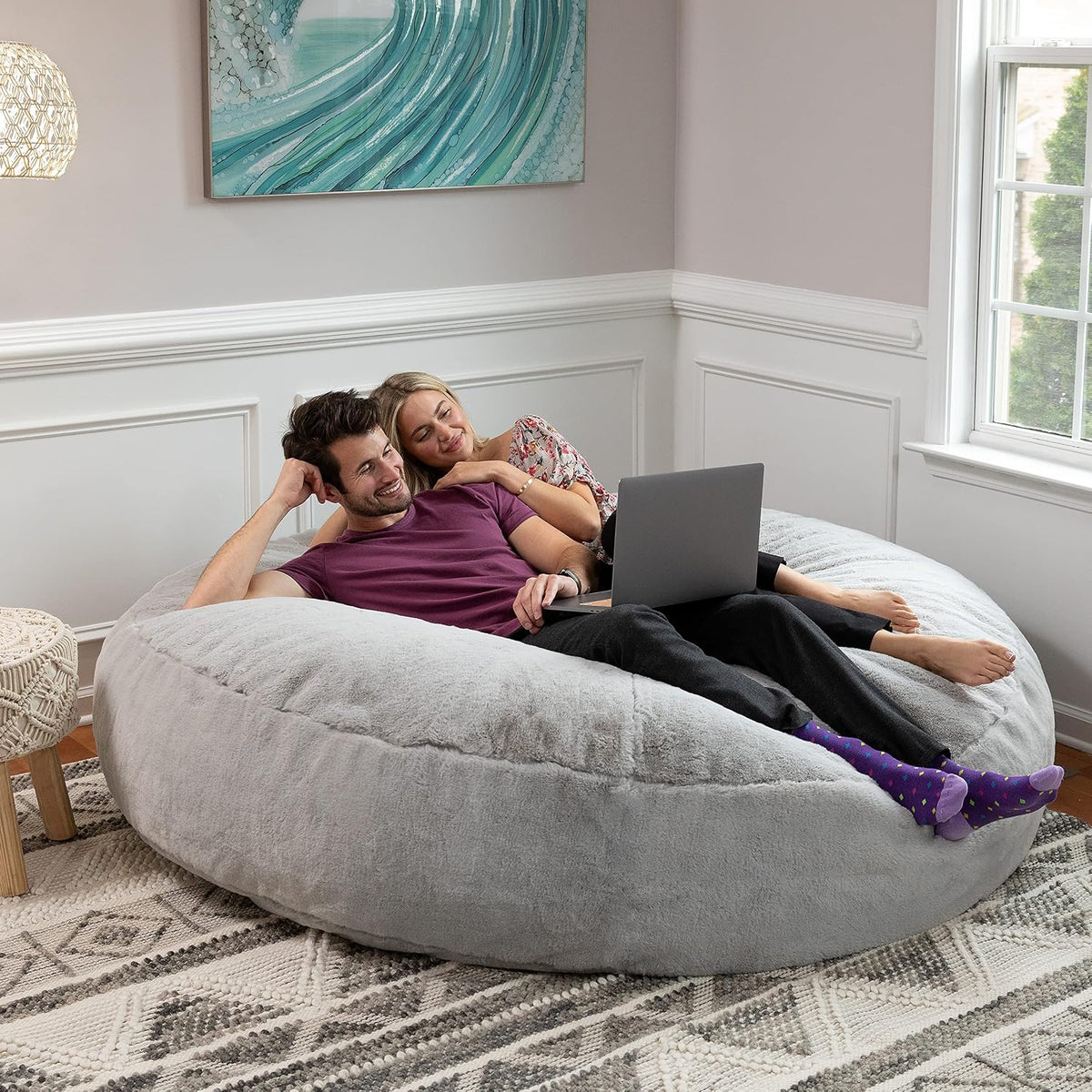 A couple lounging together on a light gray oversized bean bag while using a laptop in a bright room.