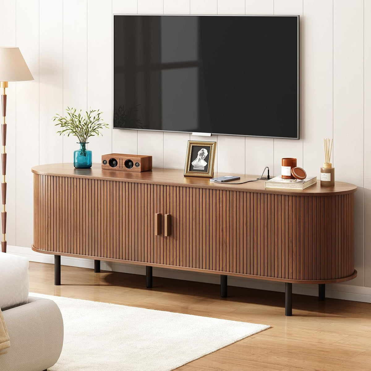 Angled walnut TV stand view with curved ends, closed tambour doors and black legs in a bright living room setting
