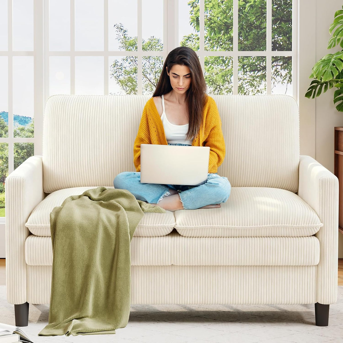 Front view of a small ribbed beige loveseat with a woman on it using a laptop and a green throw draped over the seat.