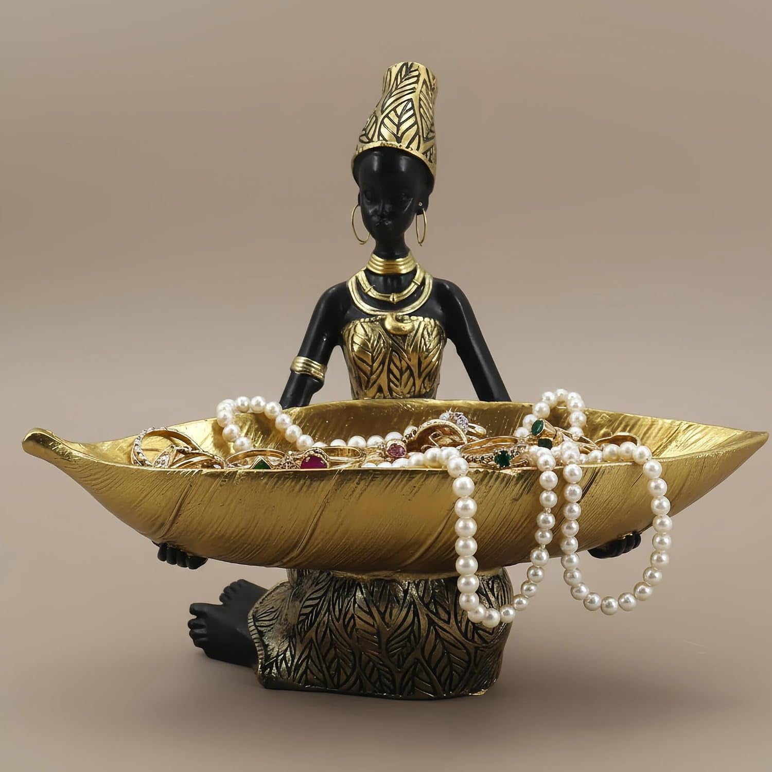 Close-up of a seated African woman figurine holding a textured gold leaf bowl filled with pearl necklaces and rings.