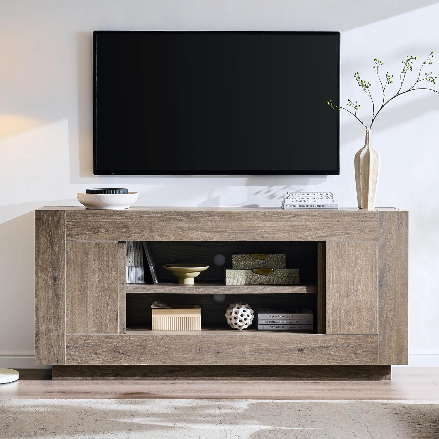 Low-profile TV stand in a light gray-brown wood finish under a wall-mounted TV, open center shelves with decor