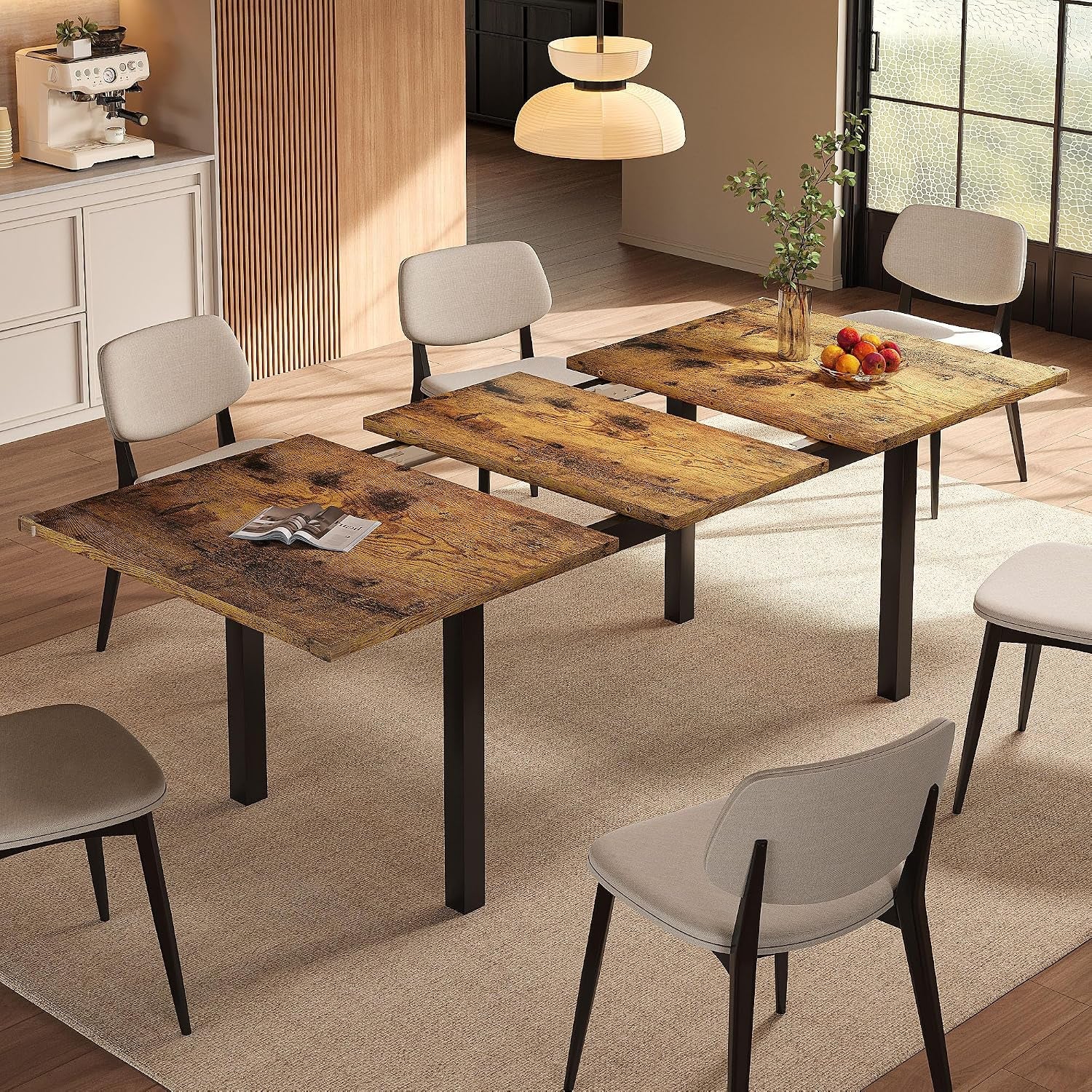 Dining room with an extendable wood‑tone table mid‑extension, black metal legs, beige upholstered chairs, vase and fruit