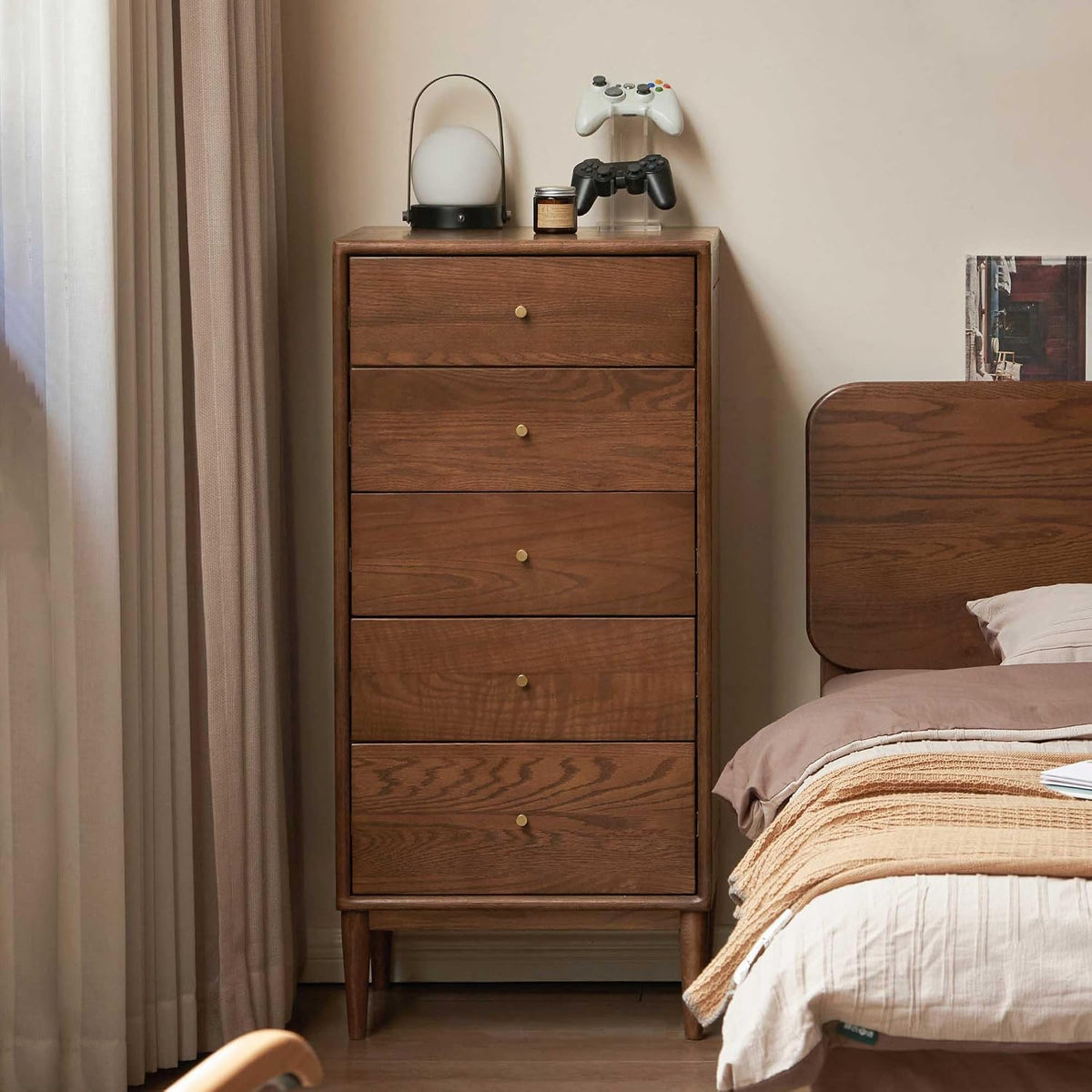 Tall dark-oak five-drawer chest beside a bed, styled with a lamp and game controllers on top.