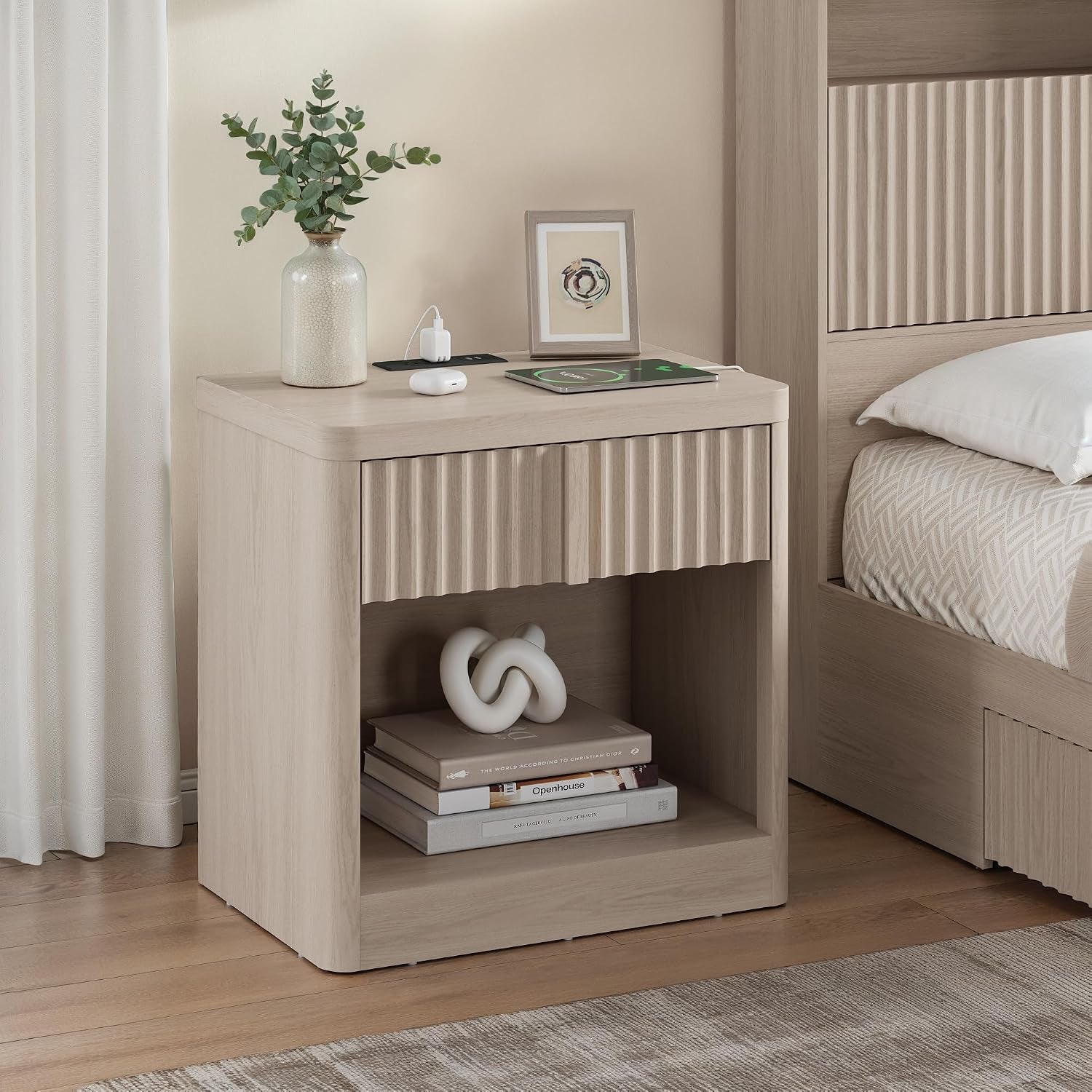 Light oak nightstand beside a bed, showing a fluted drawer top, open shelf with books, and a phone on a charger.