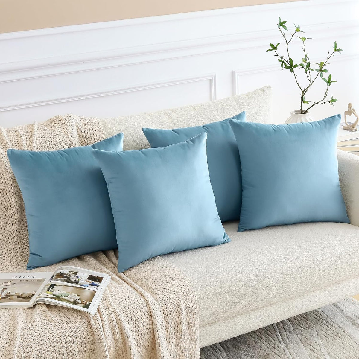 Four light-blue throw pillows arranged along a cream sofa with a small plant and open magazine nearby