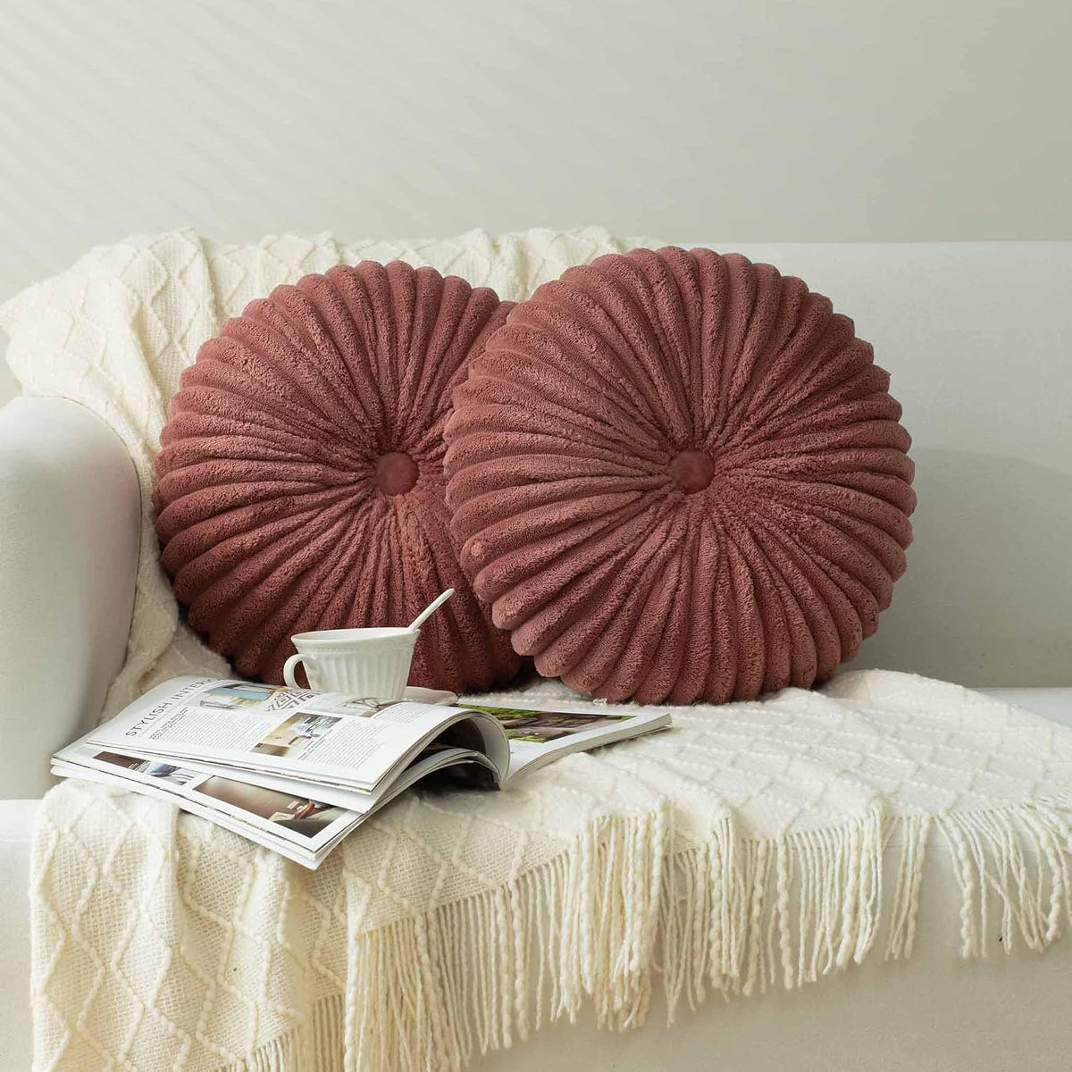 Two round dusty-rose ribbed plush pillows resting on a sofa over a textured ivory throw with a magazine.