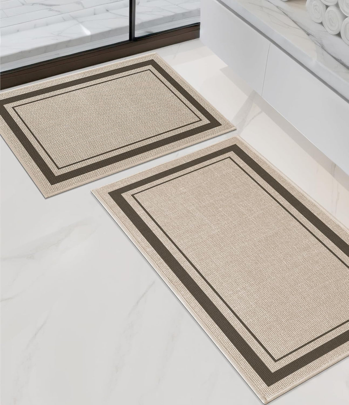 Angled bathroom view of two beige rectangular rugs with double dark-brown borders beside a white vanity.