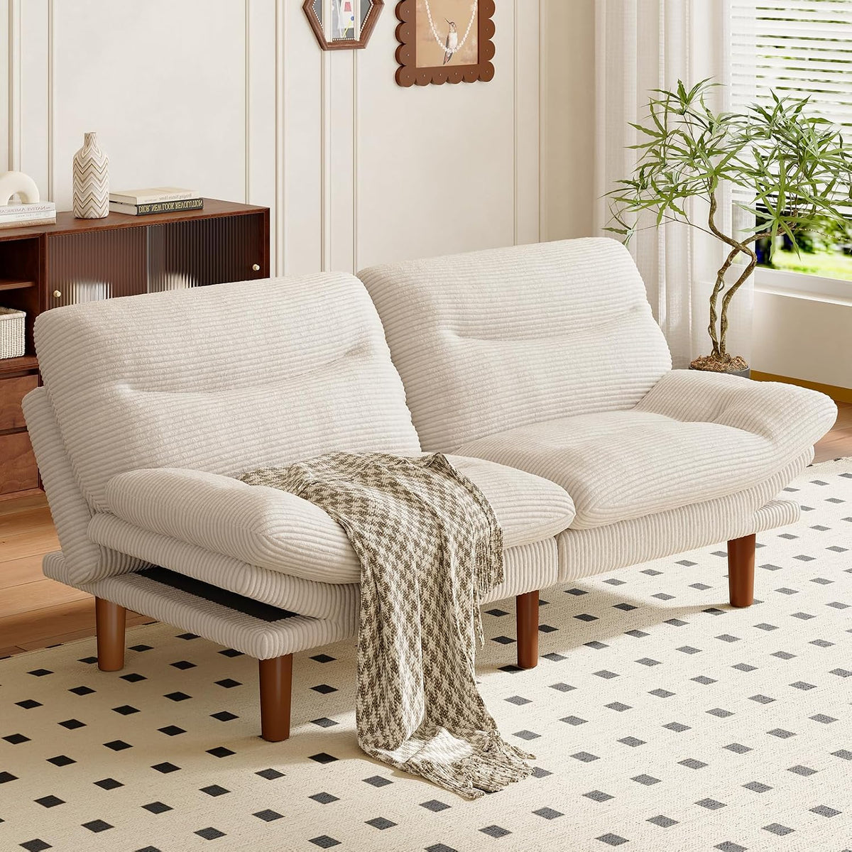 Cream corduroy convertible futon sofa with wooden legs and a patterned throw in a bright living room.
