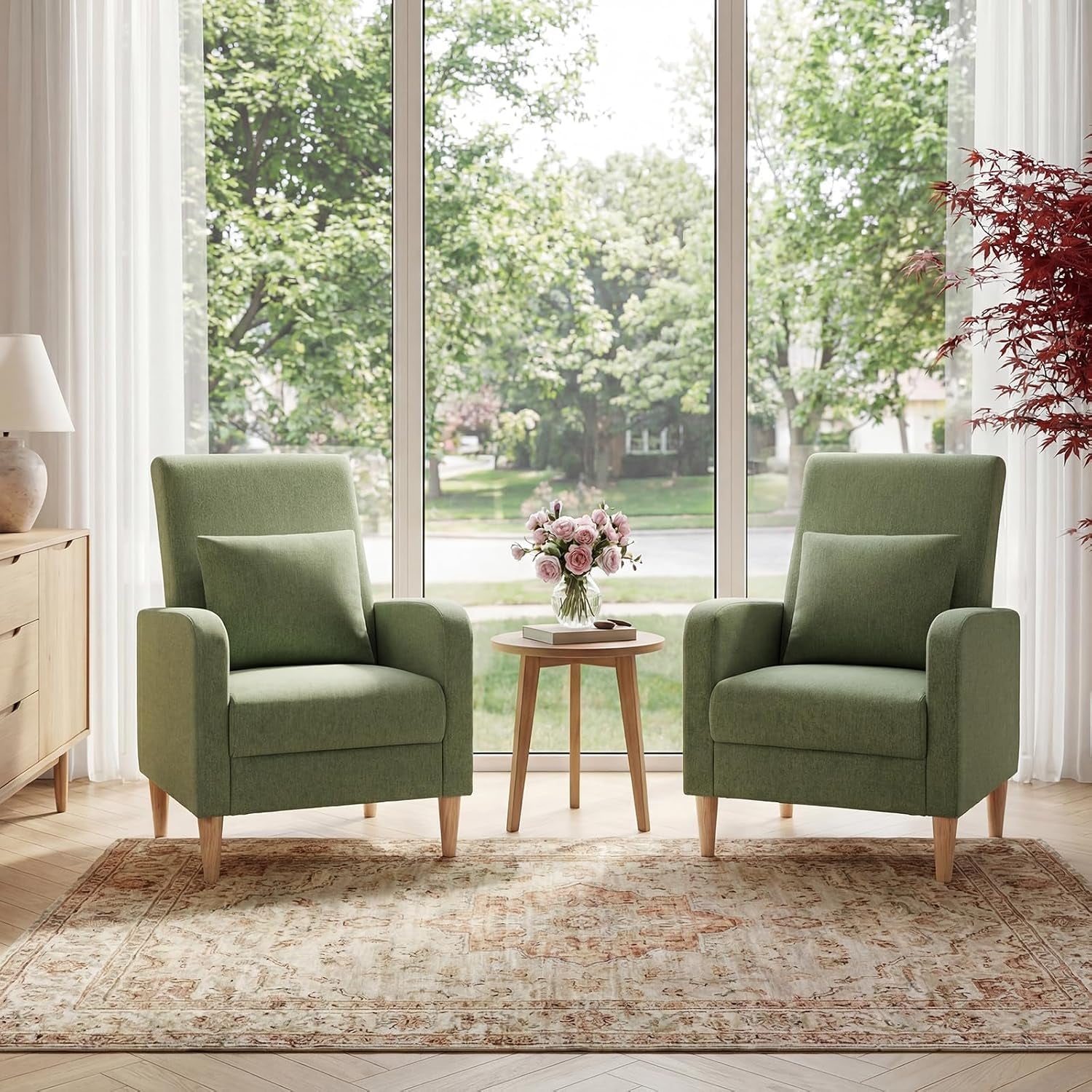 Two green fabric accent chairs flanking a small round wood table in front of tall windows