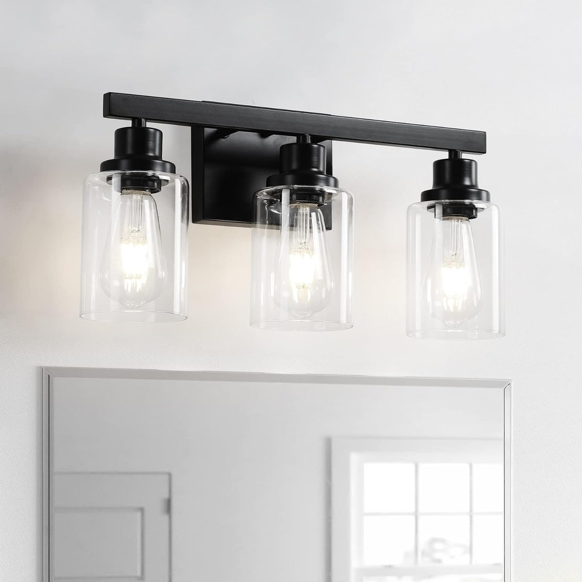 Three-light matte black vanity light with clear glass shades reflected over a bathroom mirror.