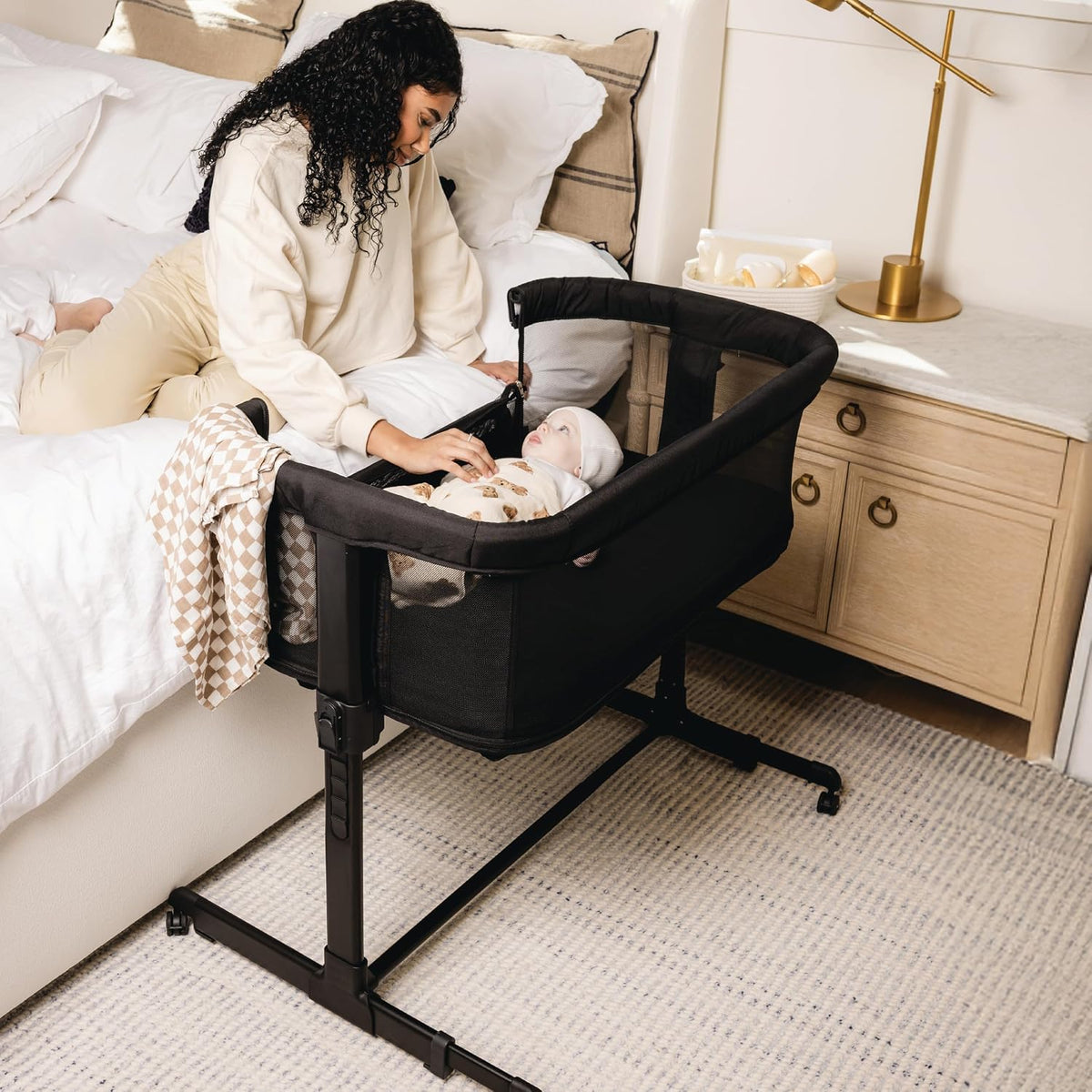 Black bedside sleeper next to a bed with a woman reaching toward an awake baby inside
