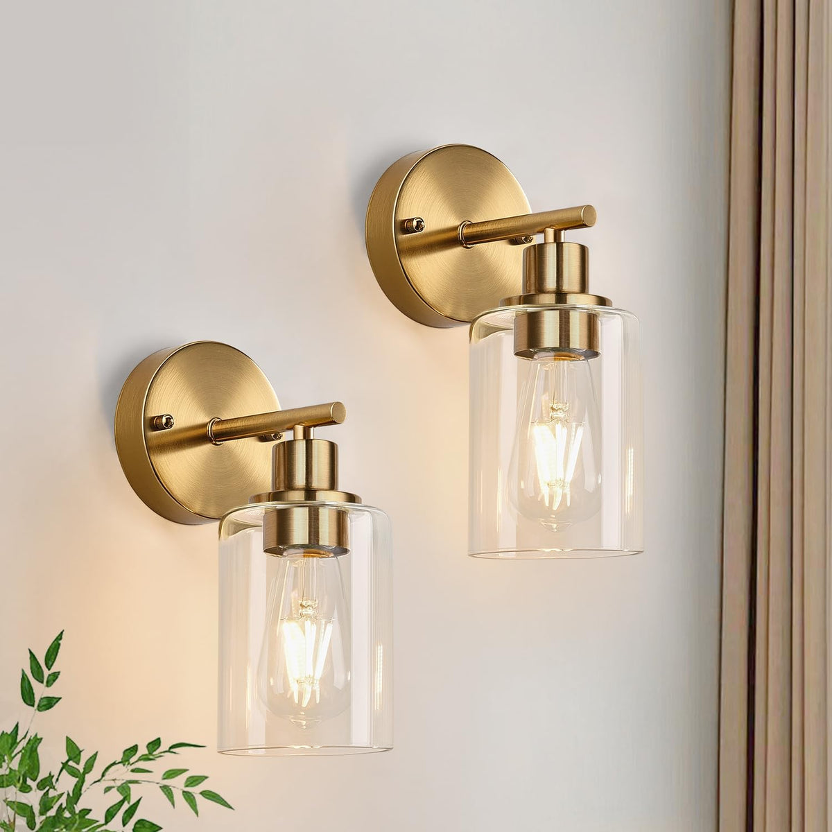 Two brushed-gold wall lights with clear glass cylinders shown in closeup on a light-colored wall.