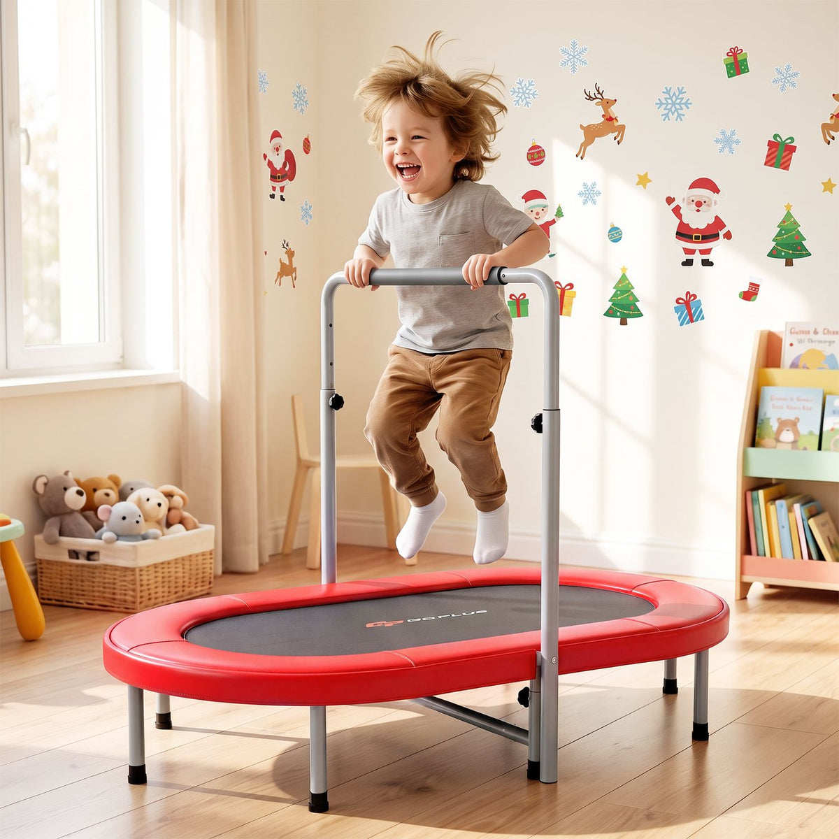 A child jumps on a red trampoline while holding its support bar in a bright playroom.