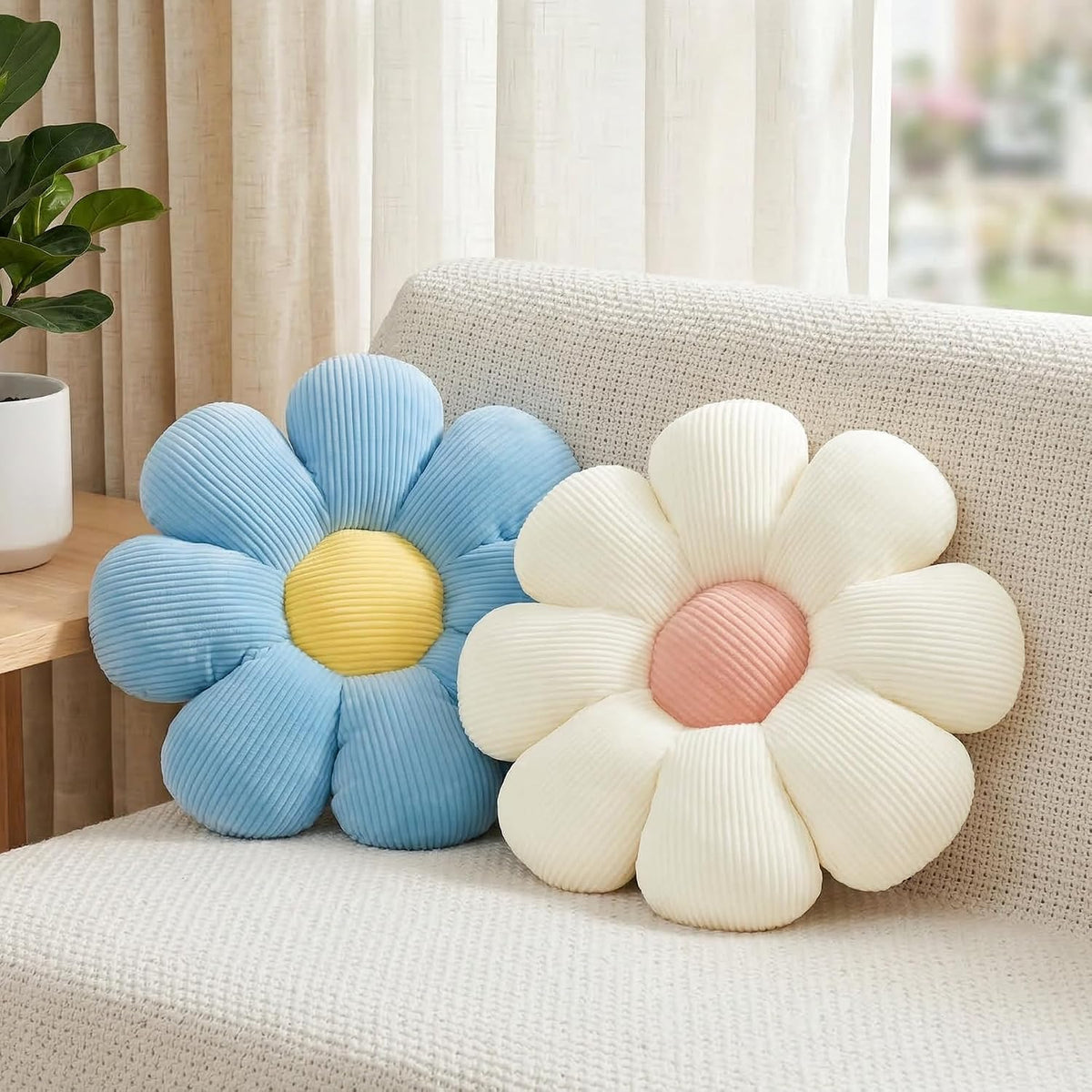 Blue daisy cushion with yellow center beside a cream daisy pillow on a textured sofa next to a potted plant.