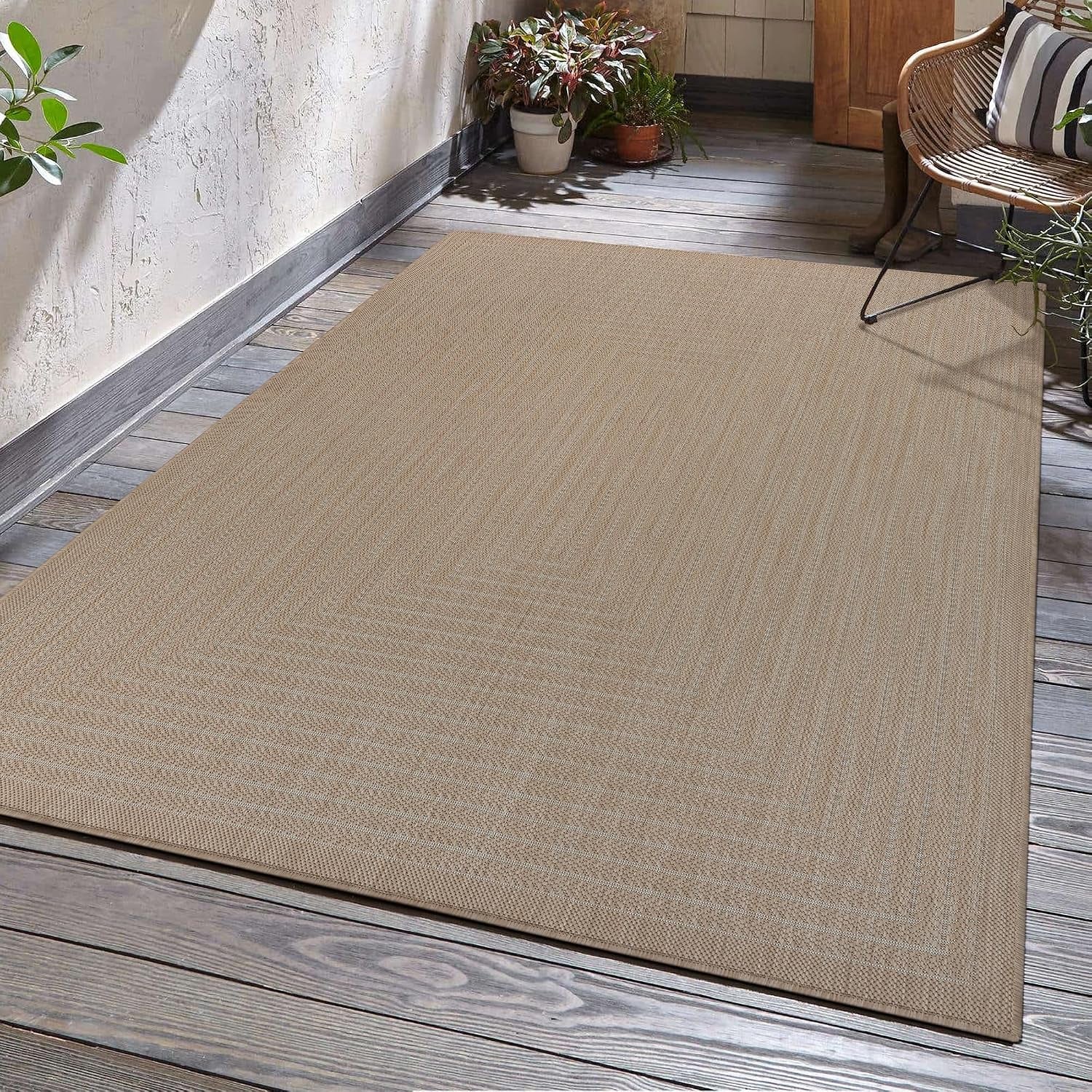Angled view of a light beige outdoor rug laid on weathered wooden deck boards beside a wall and chair.