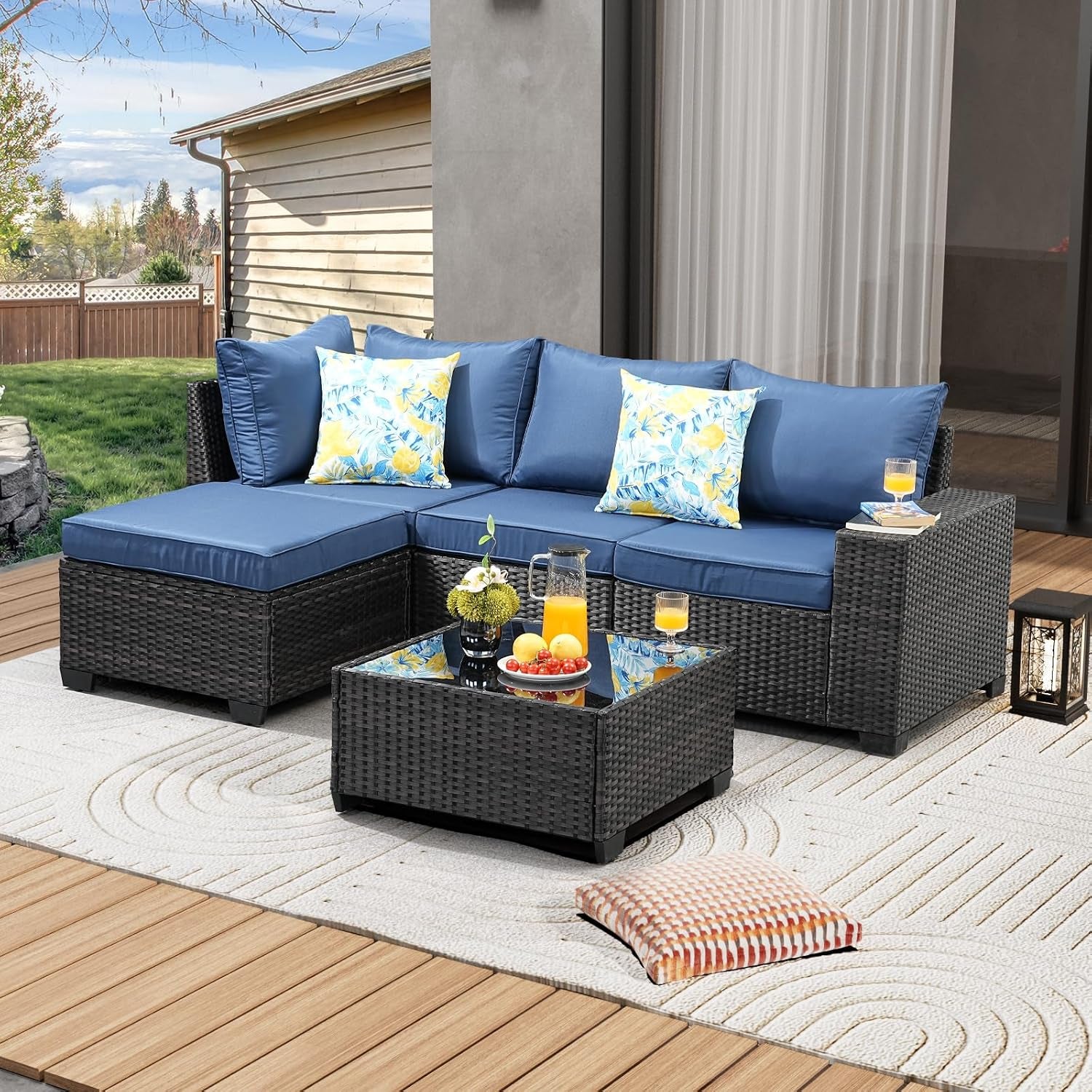 Patio sectional set with blue cushions, floral throw pillows and glass coffee table on an outdoor rug.