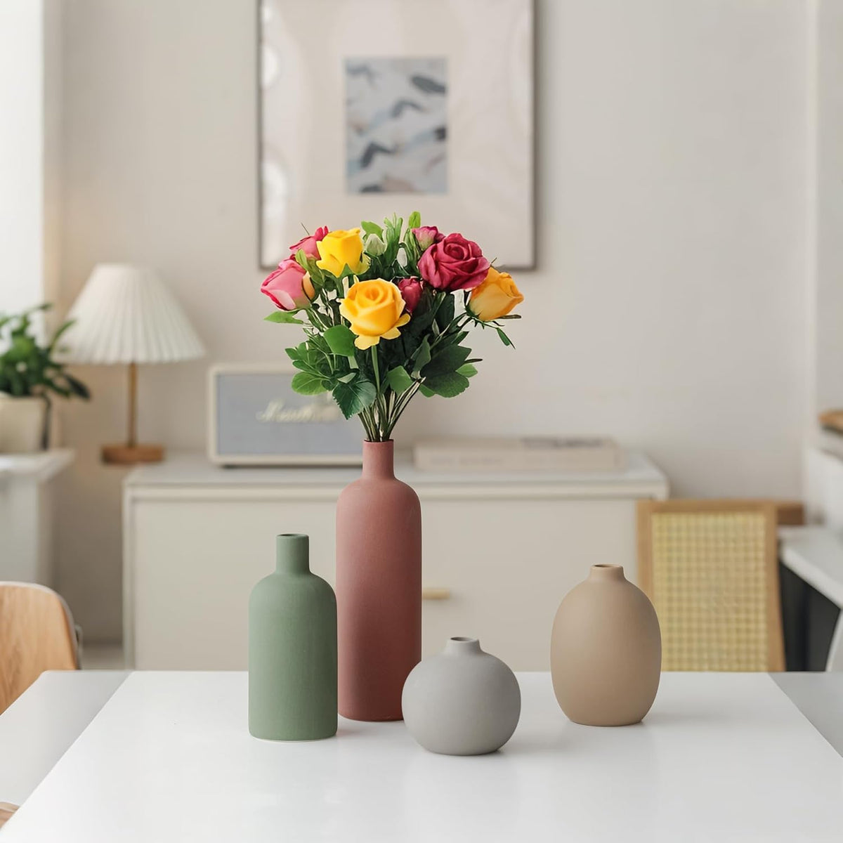 Three matte vases in green, terracotta and grey on a table with a framed print and lamp in the background
