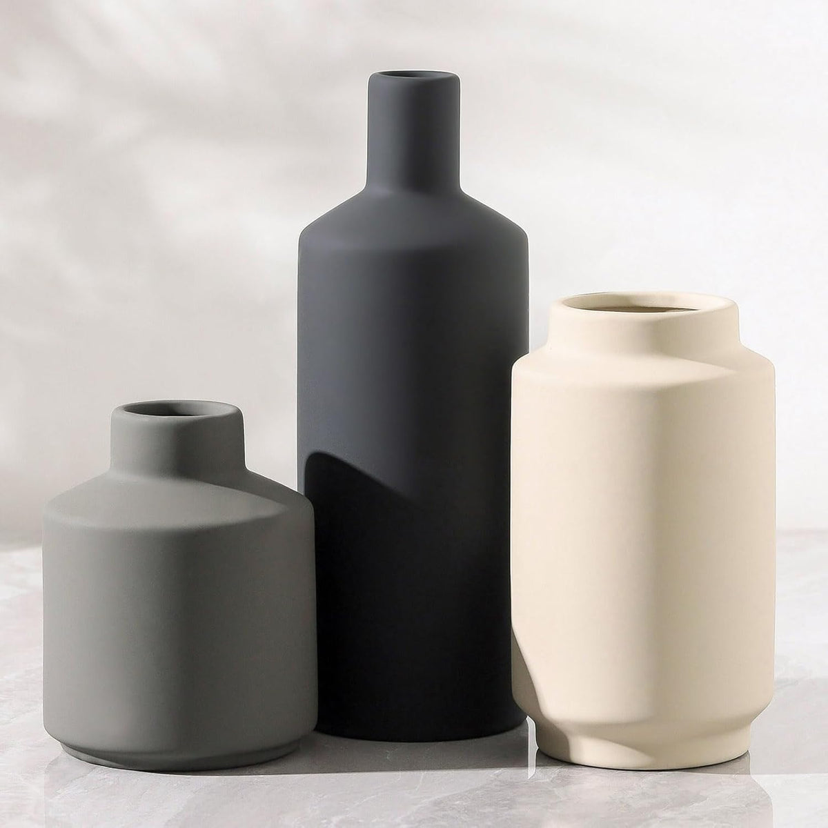 Close-up of three matte ceramic vases in grey, black and cream against a soft white backdrop, cropped vertical view