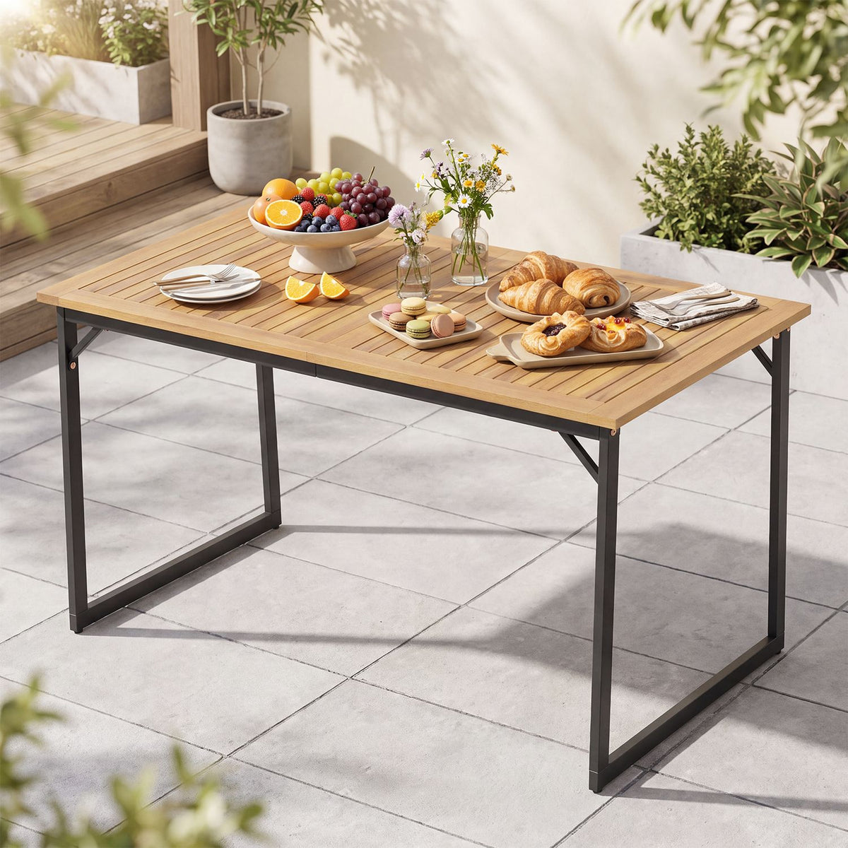 Slatted wooden dining table set with plates, fruit and pastries on a tiled patio.