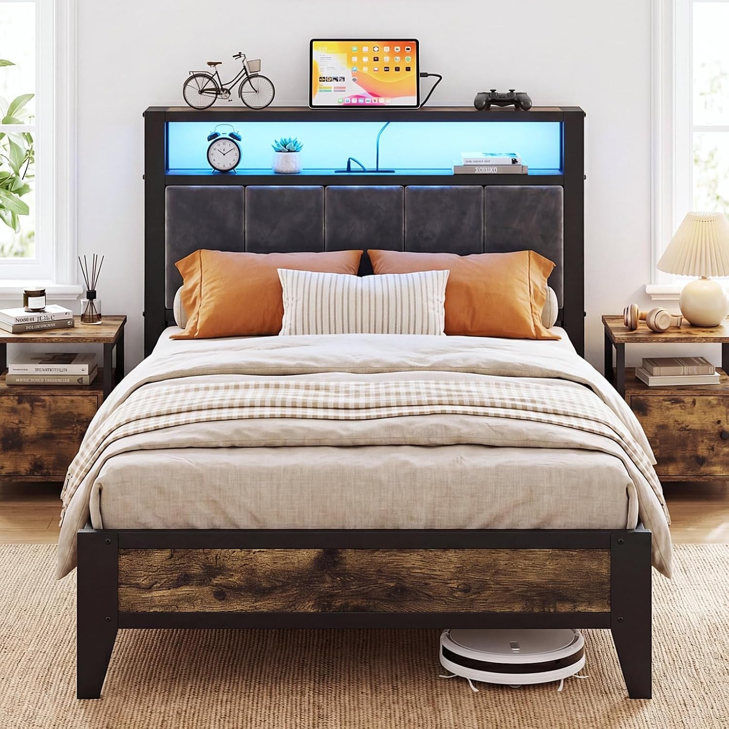 Front view of bed with blue-lit headboard shelf, gray upholstered panels, orange pillows and wood nightstands; robot vacuum
