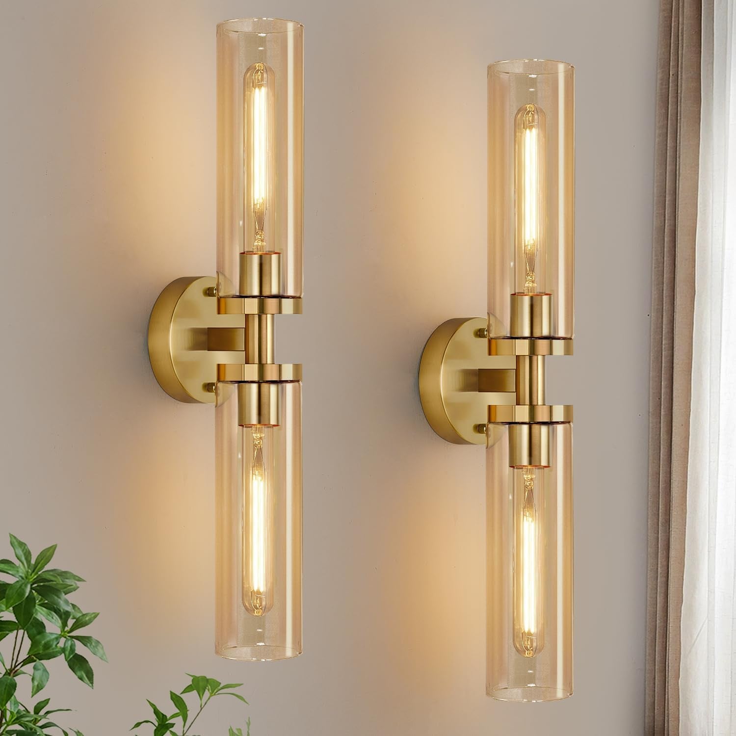 Two vertical polished-gold wall sconces with warm amber glass tubes lit on a pale wall next to a curtain.