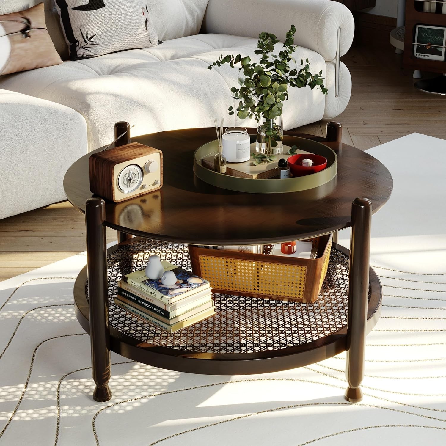 Round coffee table with darker wood finish, rounded legs and woven lower shelf, tray and decor on top in a sunlit room.