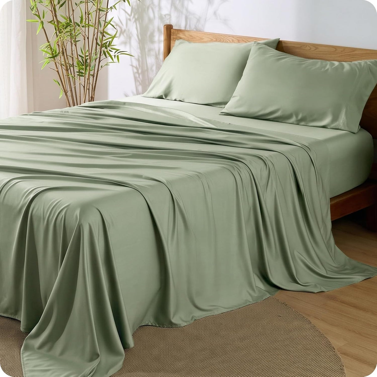 Soft sage-green sheets and two pillows on a wooden bed frame with bamboo plant at the side.