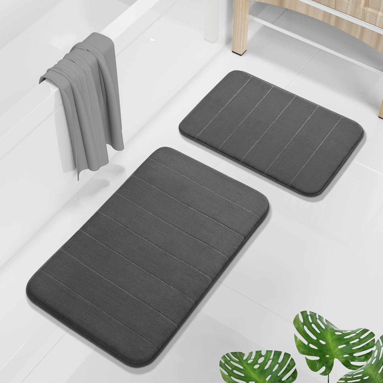Two dark gray memory-foam bathroom mats on white tile next to a bathtub and wooden cabinet
