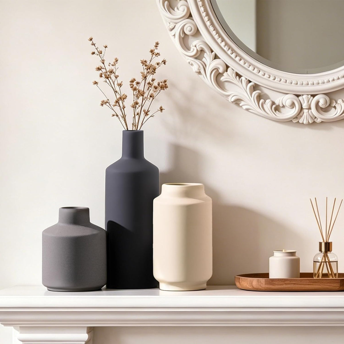 Three vases lined on a white mantel beside an ornate round mirror and a reed diffuser