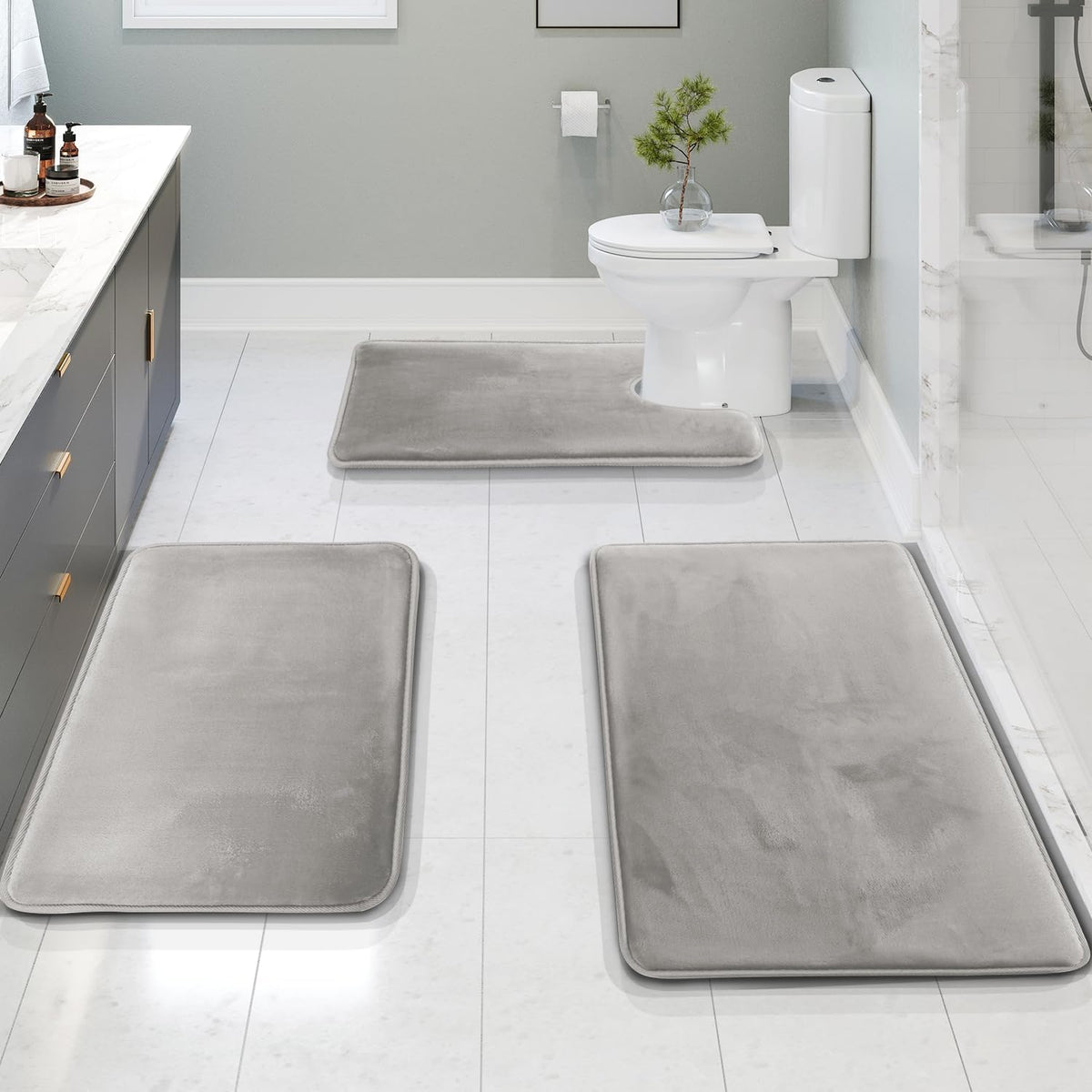 Overhead view of a three-piece light-gray bath rug set arranged on white tile: two rectangles in front and the contour piece
