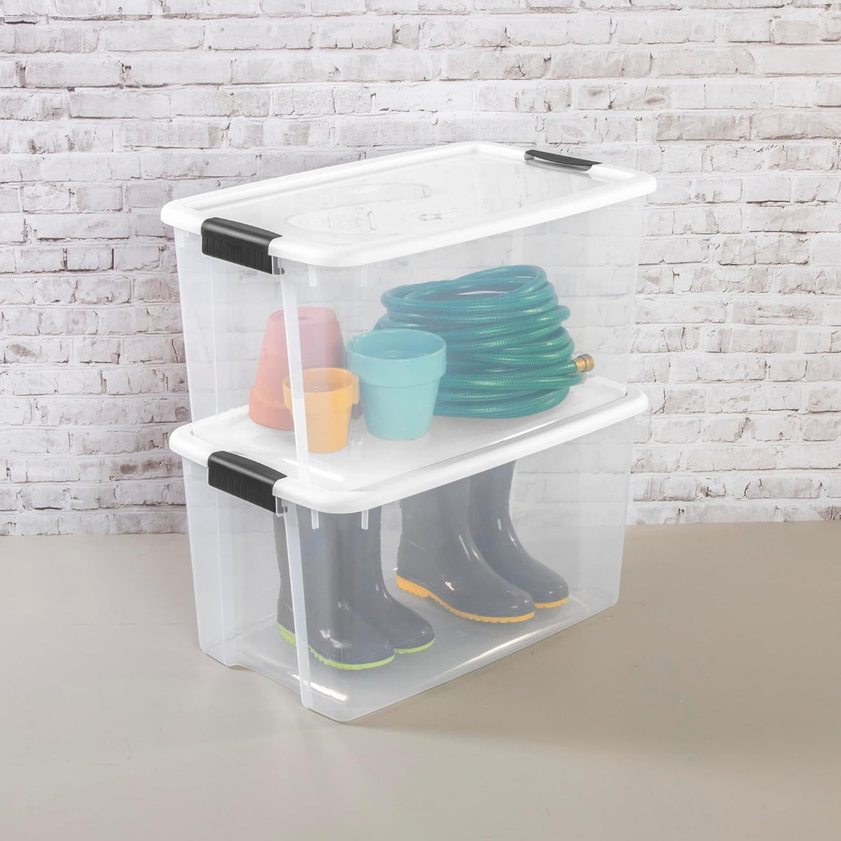 Close-up of two clear boxes stacked showing a coiled garden hose, plant pots and tall rain boots inside.