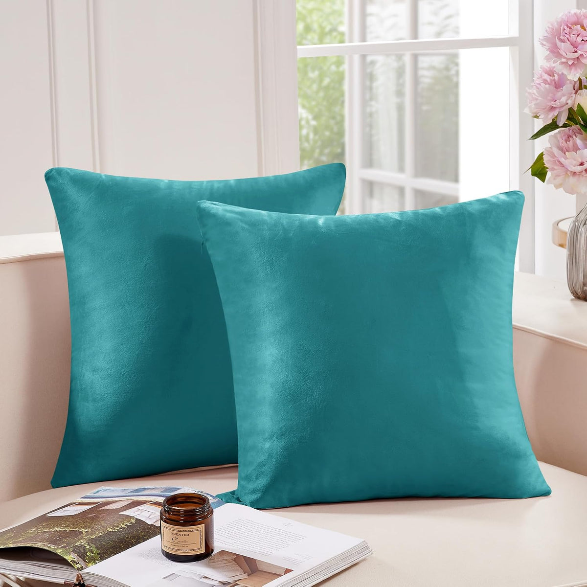 Two square teal pillow covers displayed on a sofa by a window with flowers nearby