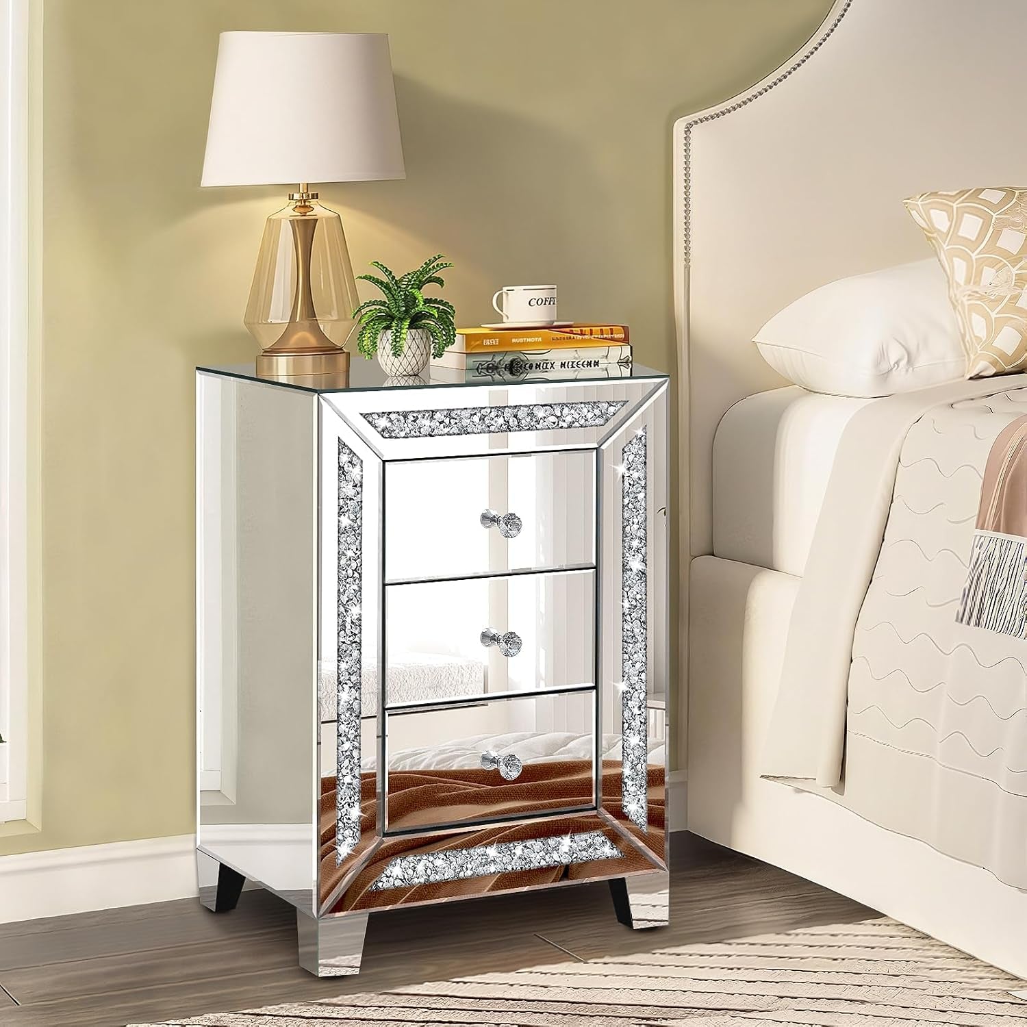 Mirrored nightstand next to a bed topped with a lamp, small potted plant, stacked books and a coffee mug.