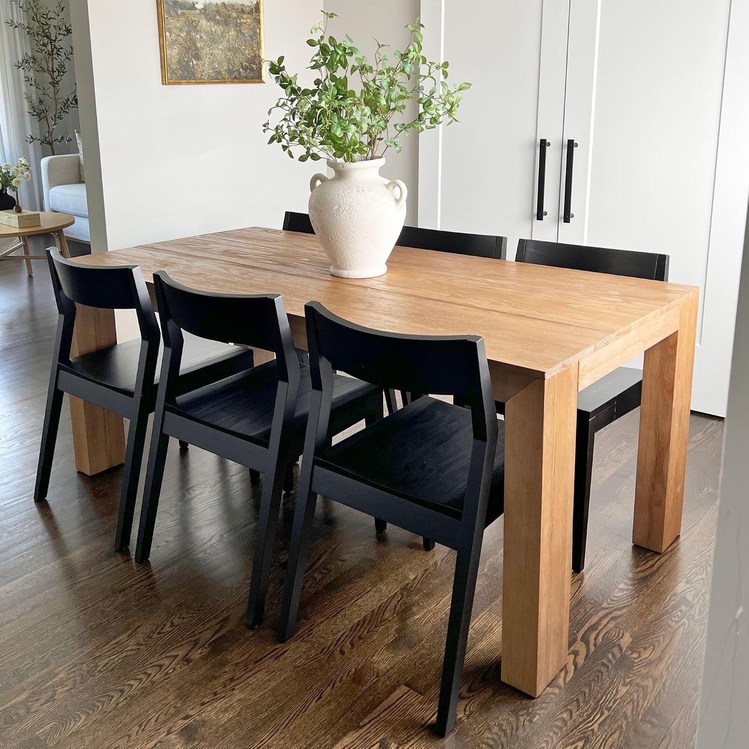 Rectangular pine dining table with six black chairs and a large vase of greenery on top in a bright room.