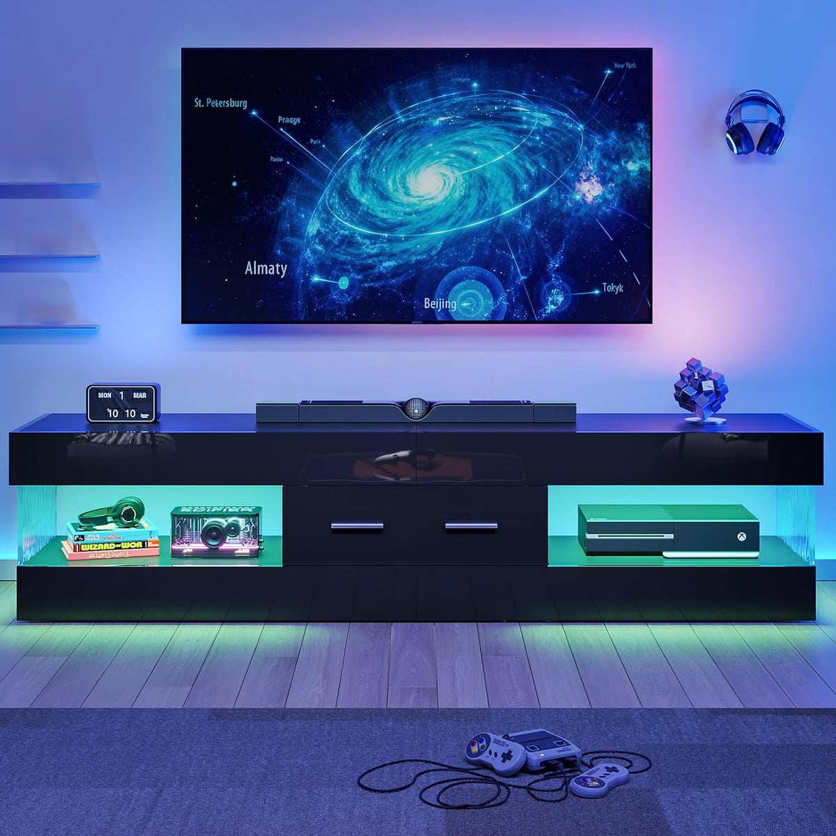 Black TV stand front view showing center drawers and green LED under-shelf lighting with game controllers on floor
