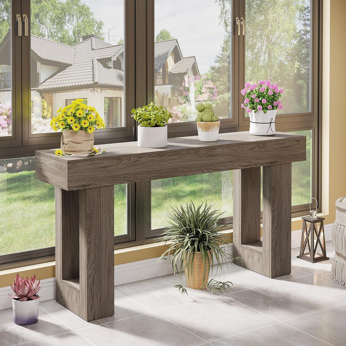 Grey wood console table beside large windows with potted plants on top and tiled floor visible