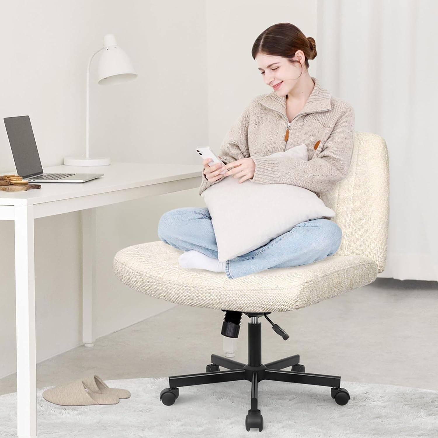 Lifestyle view of a person seated cross‑legged on a cream armless swivel chair at a light wood desk in a bright room
