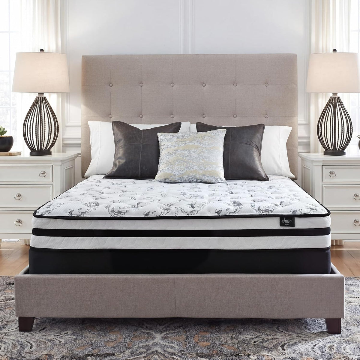 Bedroom scene with a tufted beige headboard, decorative pillows and a white floral-patterned pillow-top mattress on a fabric
