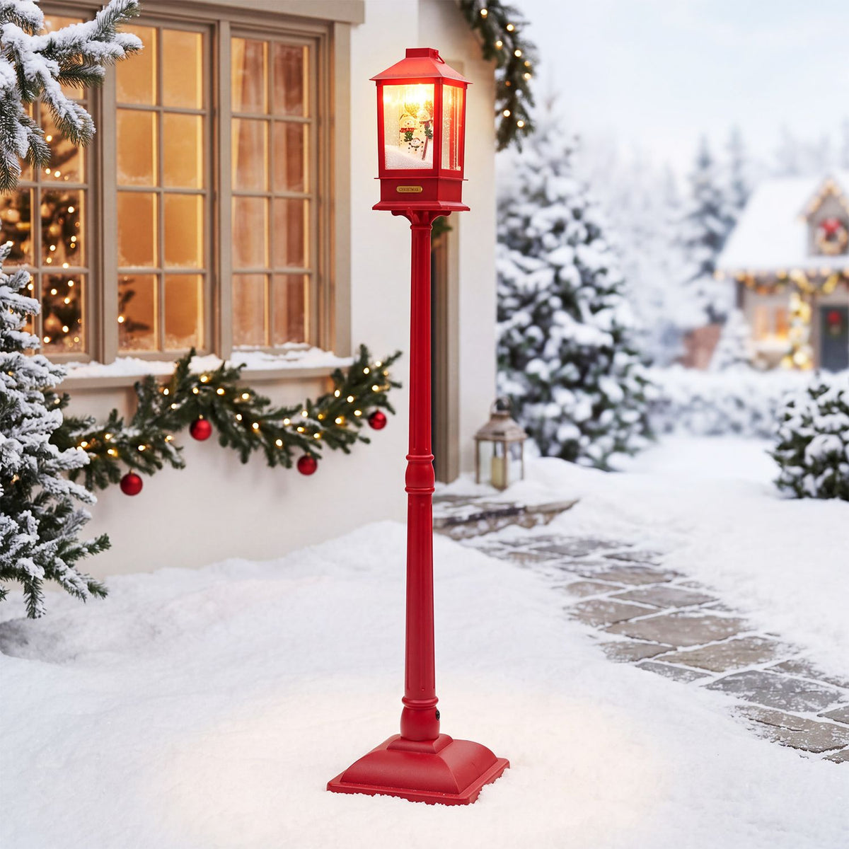 Red Christmas streetlight glowing beside a snow-covered house and decorated evergreen branches.