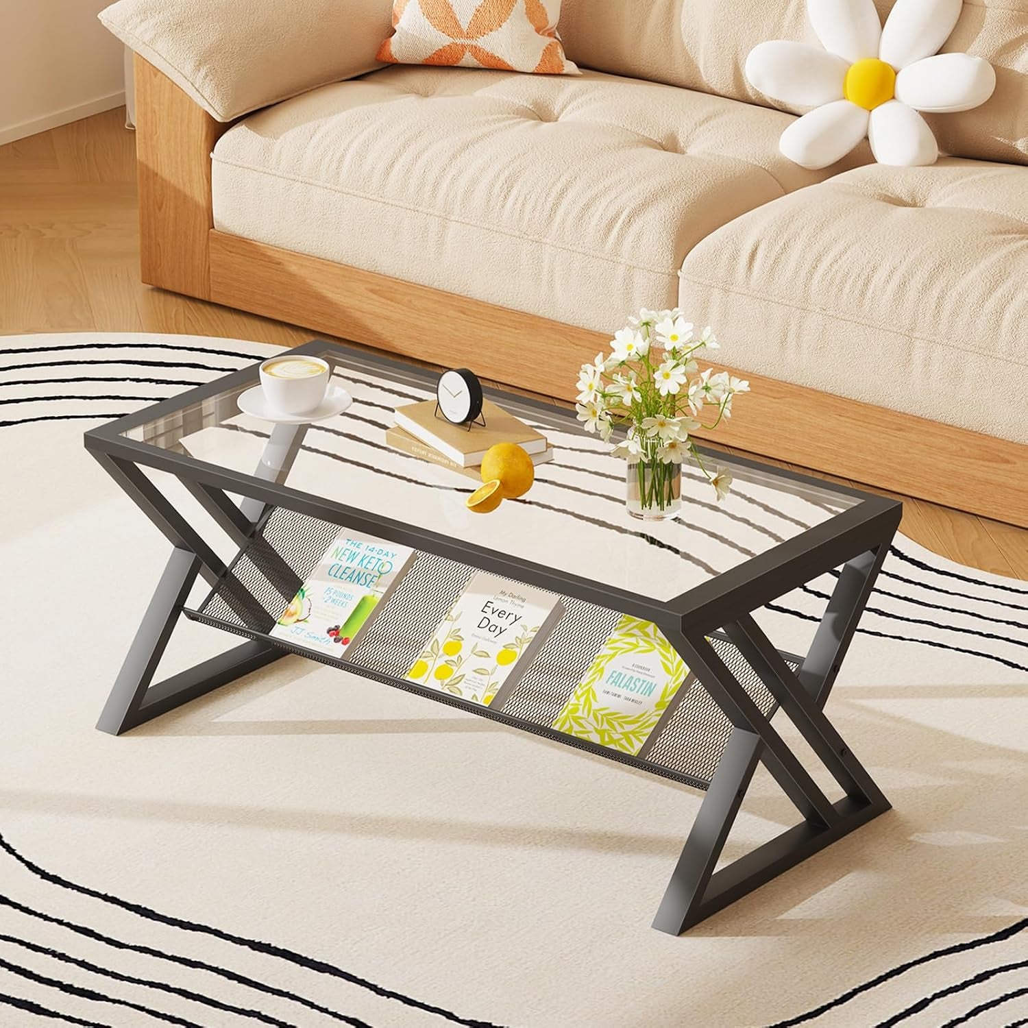 Black-framed rectangular coffee table with glass top and a mesh lower shelf holding books, placed before a beige sofa.