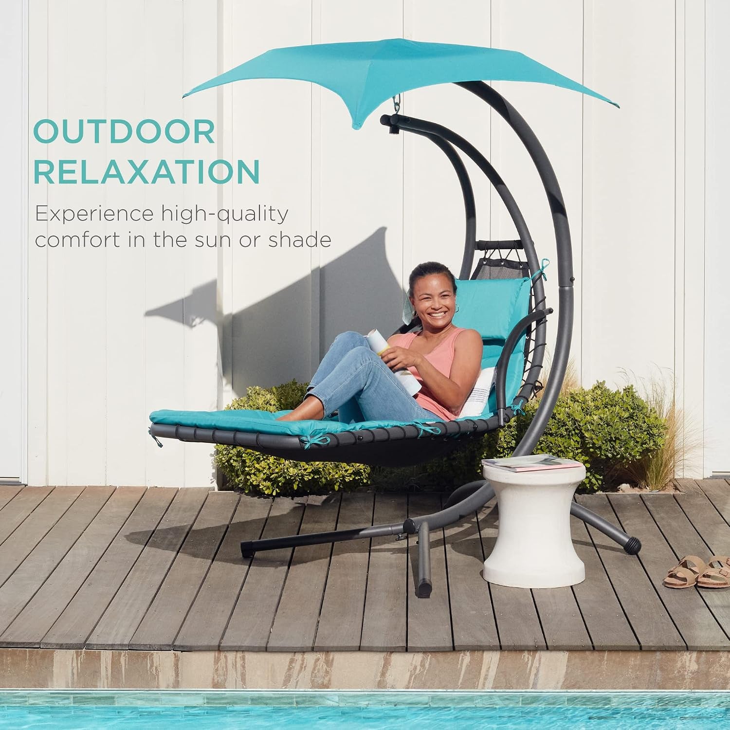 Woman relaxing on a teal hanging chaise with canopy on a wooden poolside deck beside a small table and pool.