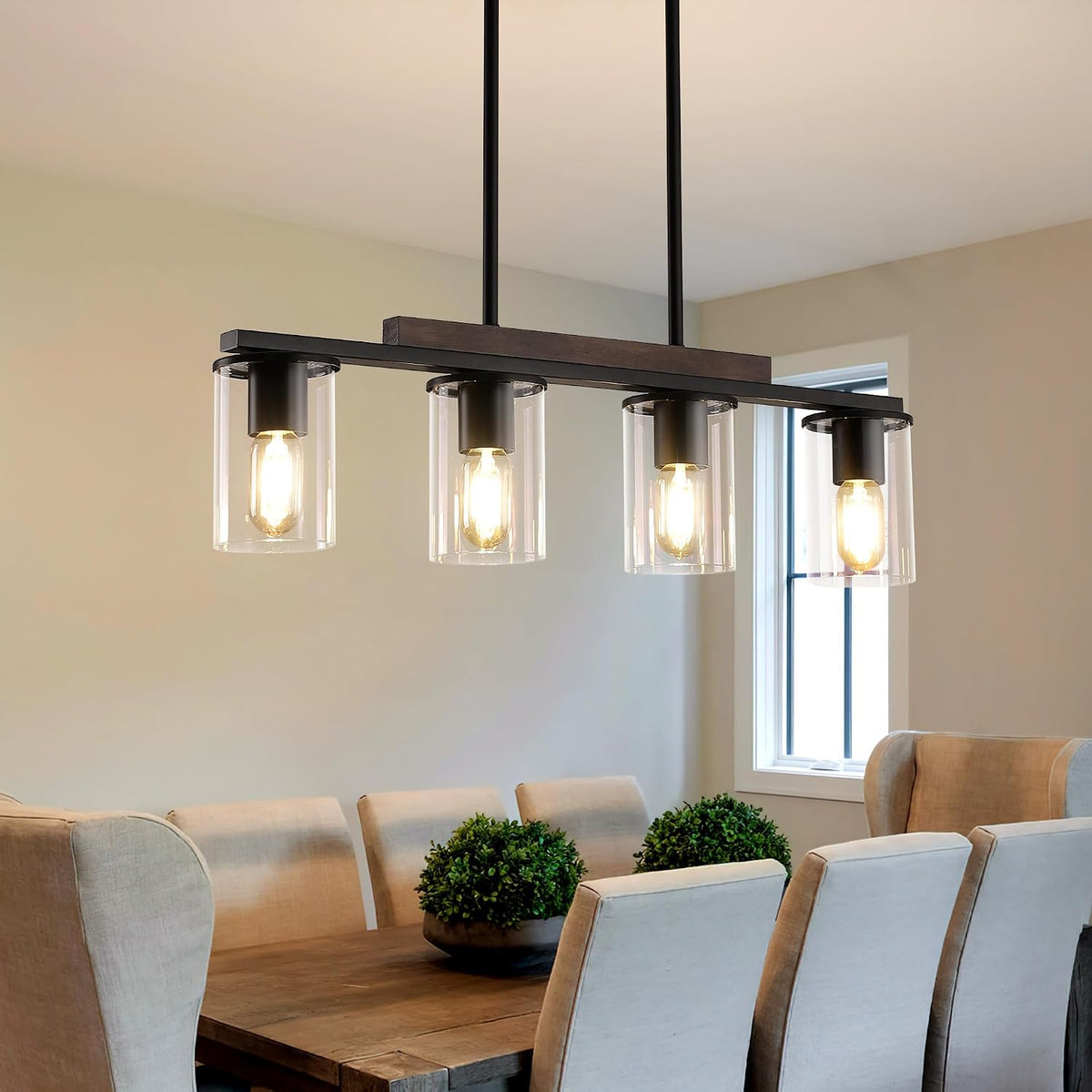 Linear chandelier lit above a wooden dining table with upholstered chairs and small greenery centerpieces.