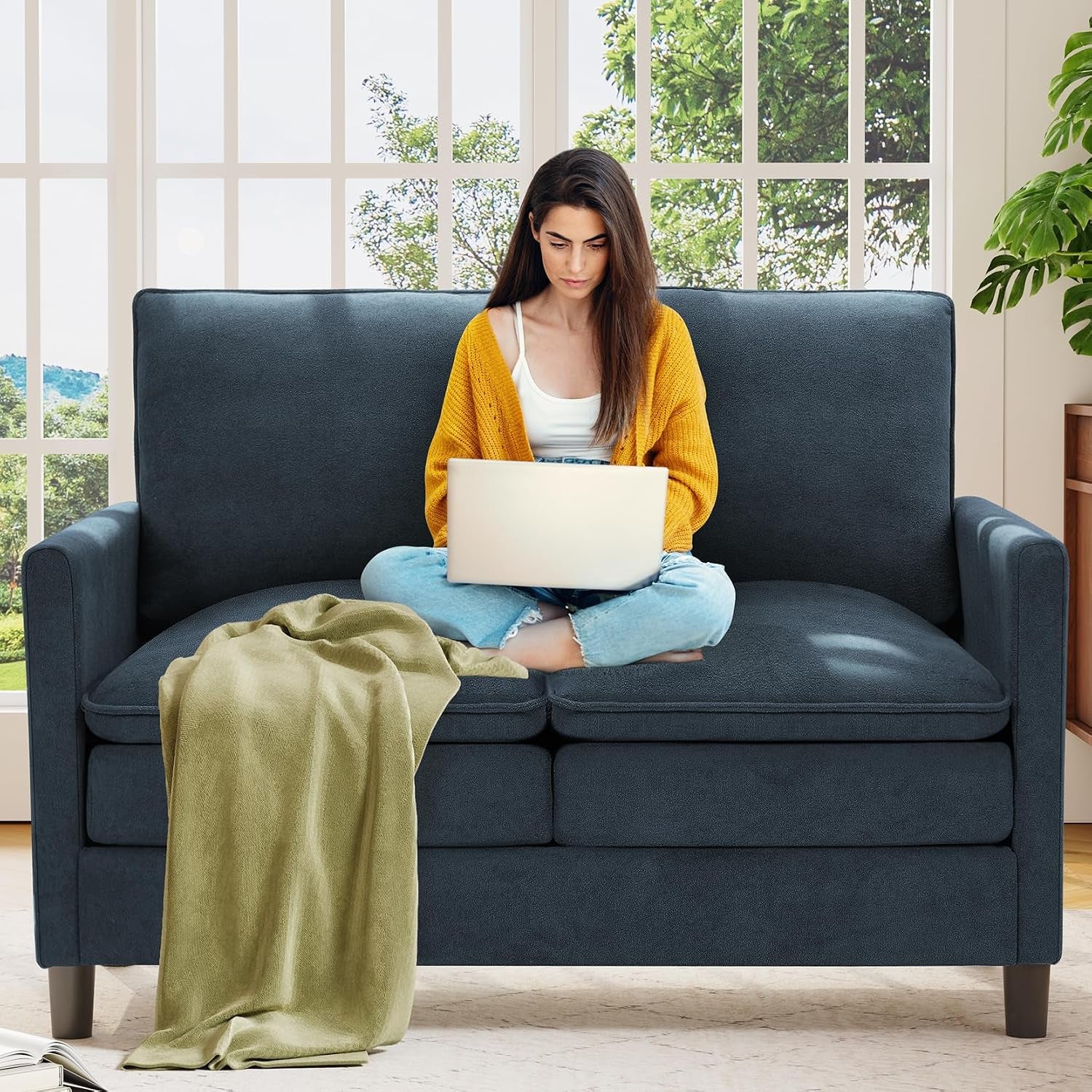 Woman working on a laptop on a smooth dark blue loveseat with an olive throw draped over the cushions.