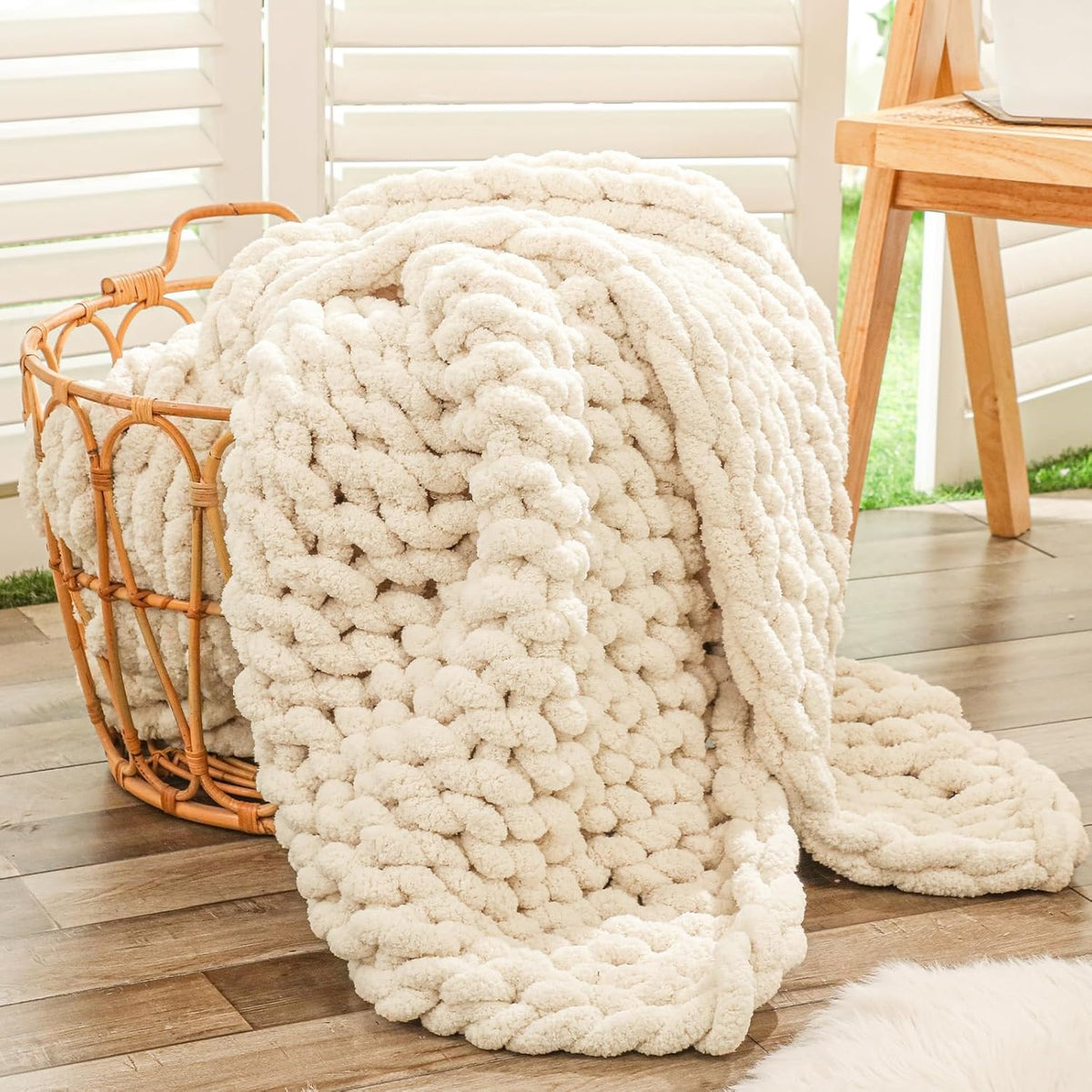 Beige chunky knit throw spilling from a woven basket on a wood floor by shuttered windows.